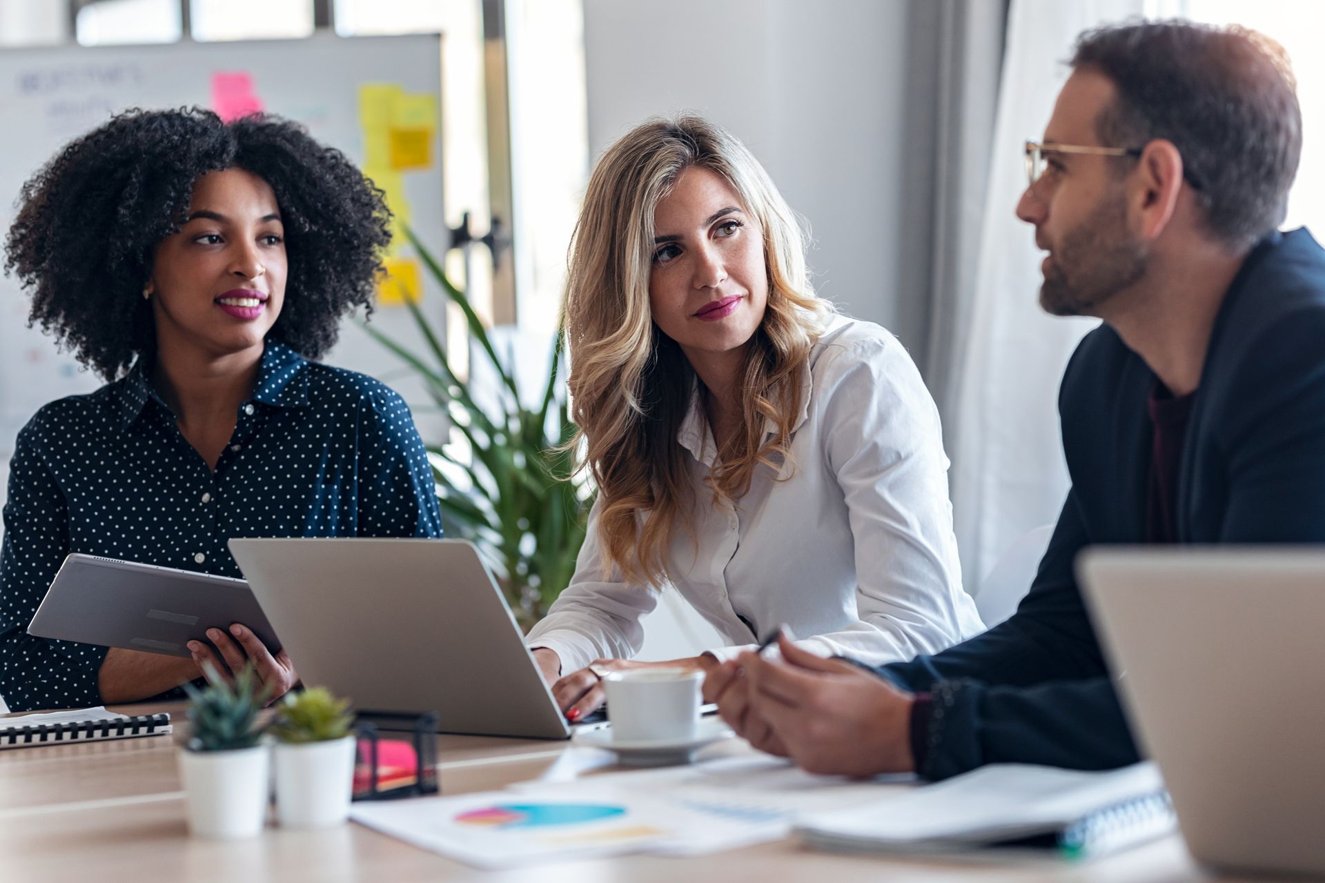 Three coworkers in a meeting, discussing documents around a laptop in a bright office.