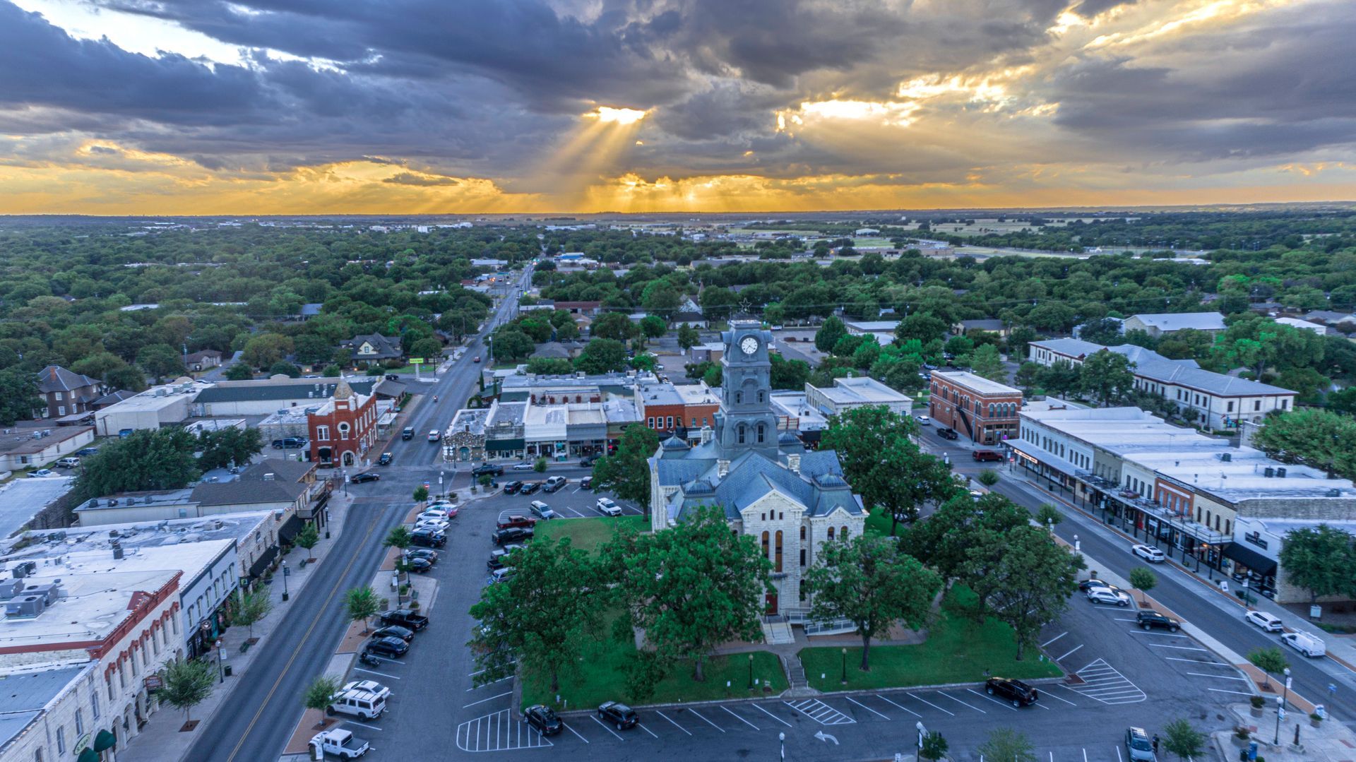 Aerial view of a small town square featuring a central historic courthouse, surrounded by streets, trees, and sunset.