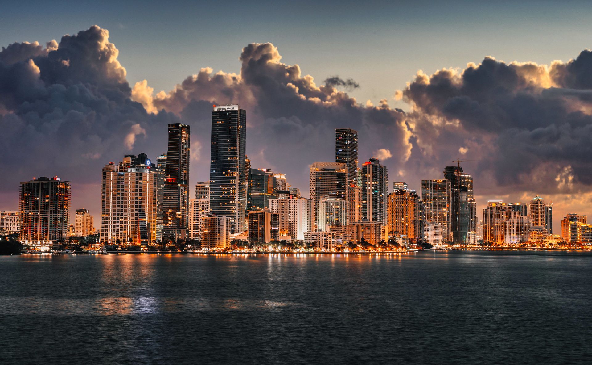 City skyline at dusk with glowing high-rise towers, dramatic clouds, and dark water in the foreground