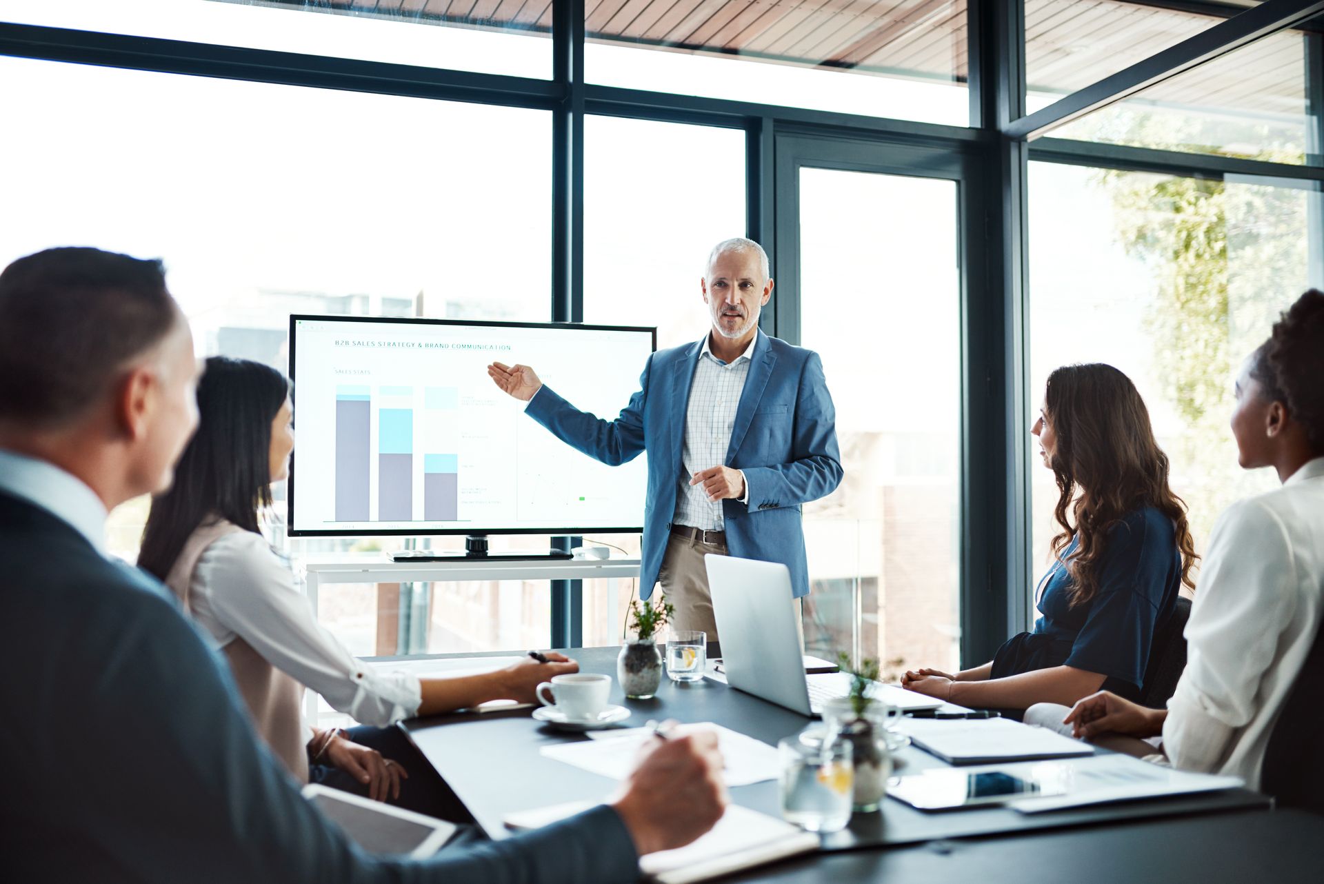 Business meeting with a presenter pointing at a chart on a screen while colleagues watch around a conference table