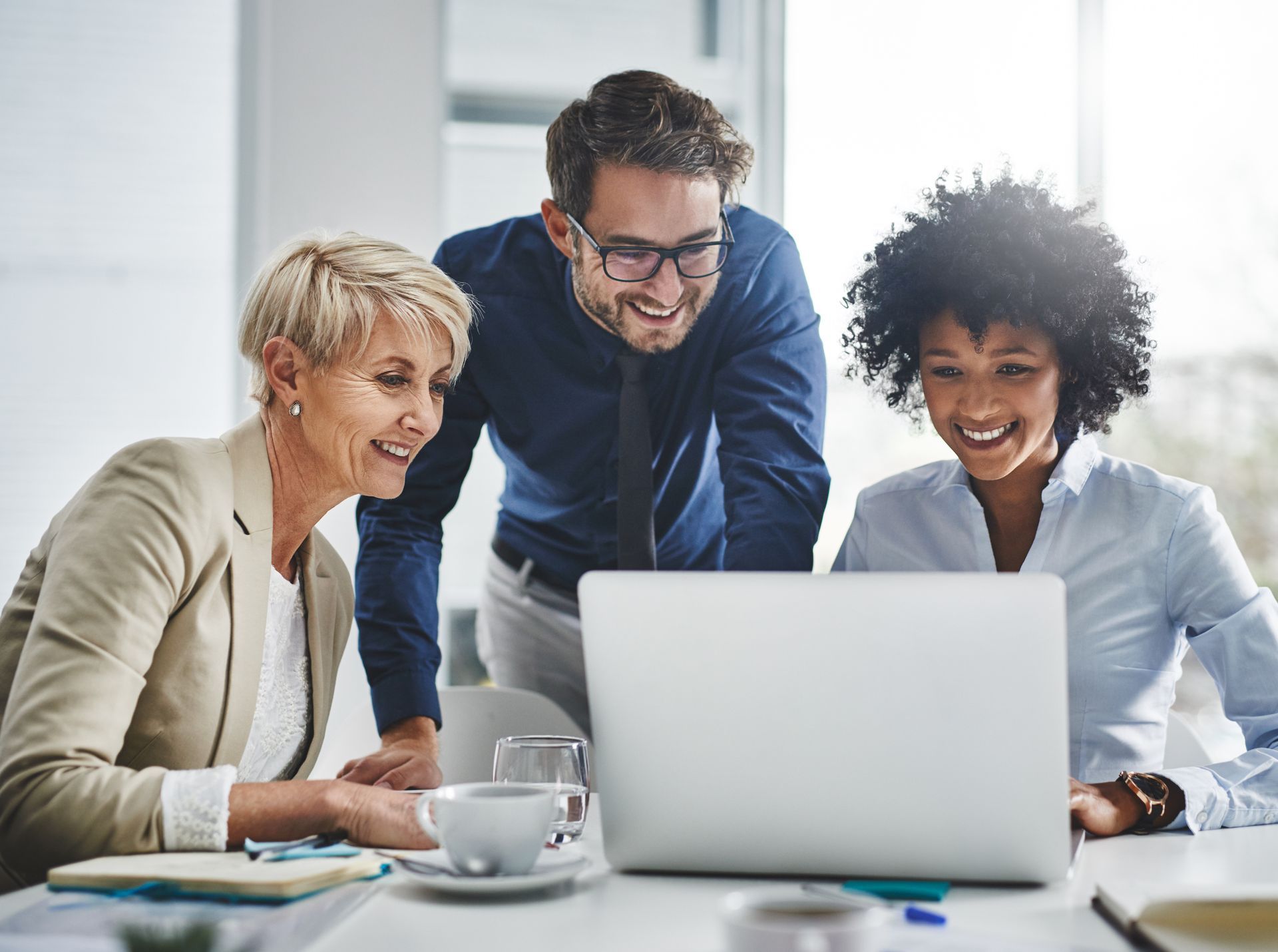 Three coworkers smiling around a laptop in a bright office, reviewing work together
