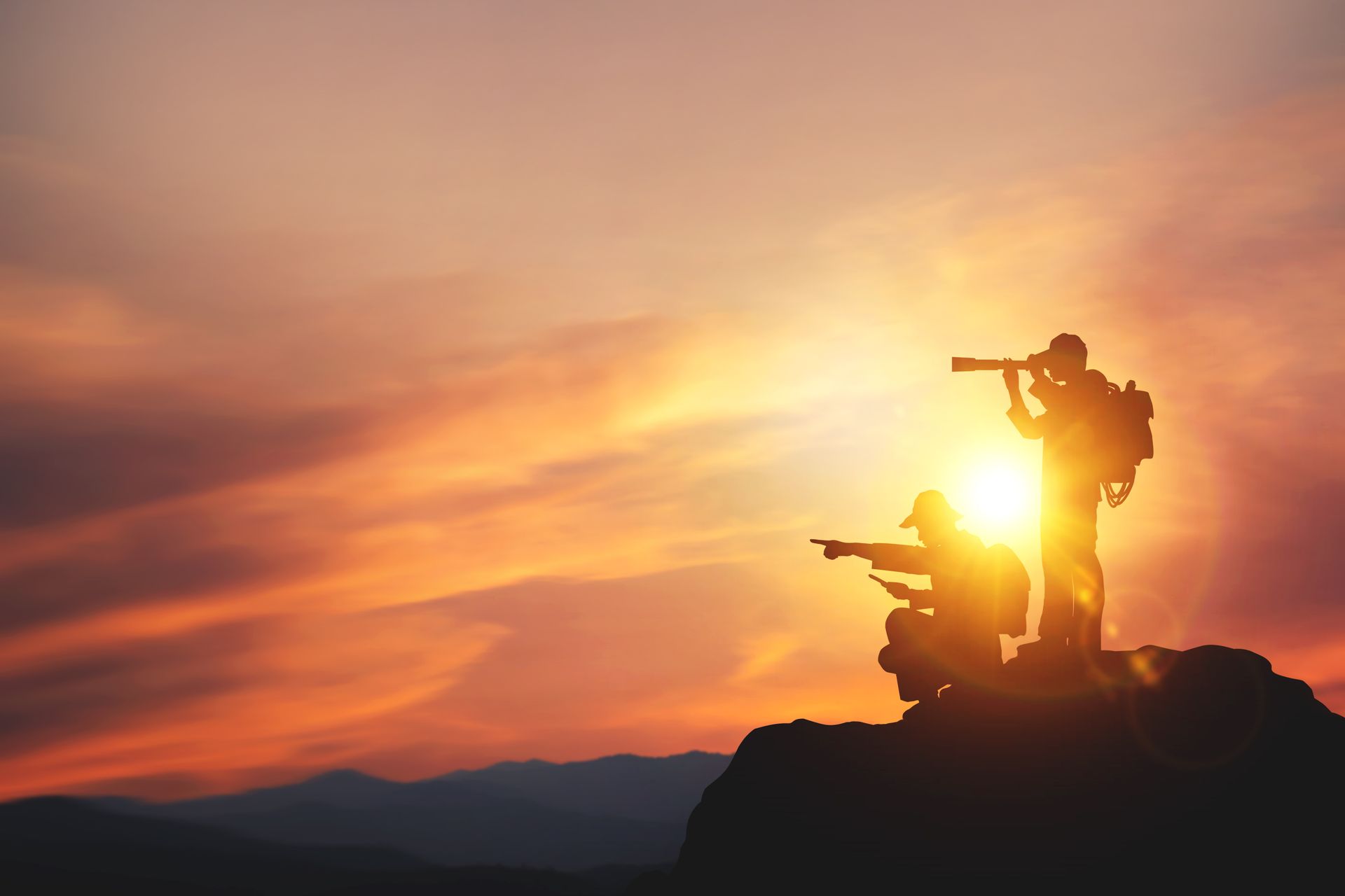 Silhouetted people on a mountain peak at sunset, one using binoculars, others pointing.