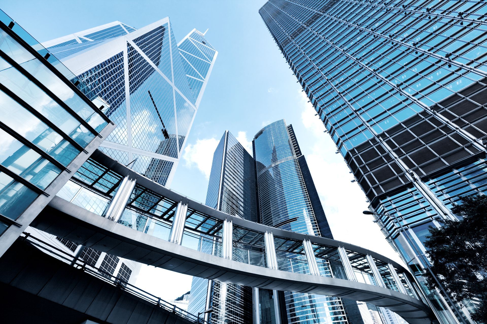 Looking up at modern glass skyscrapers and a curved pedestrian skybridge against a blue sky