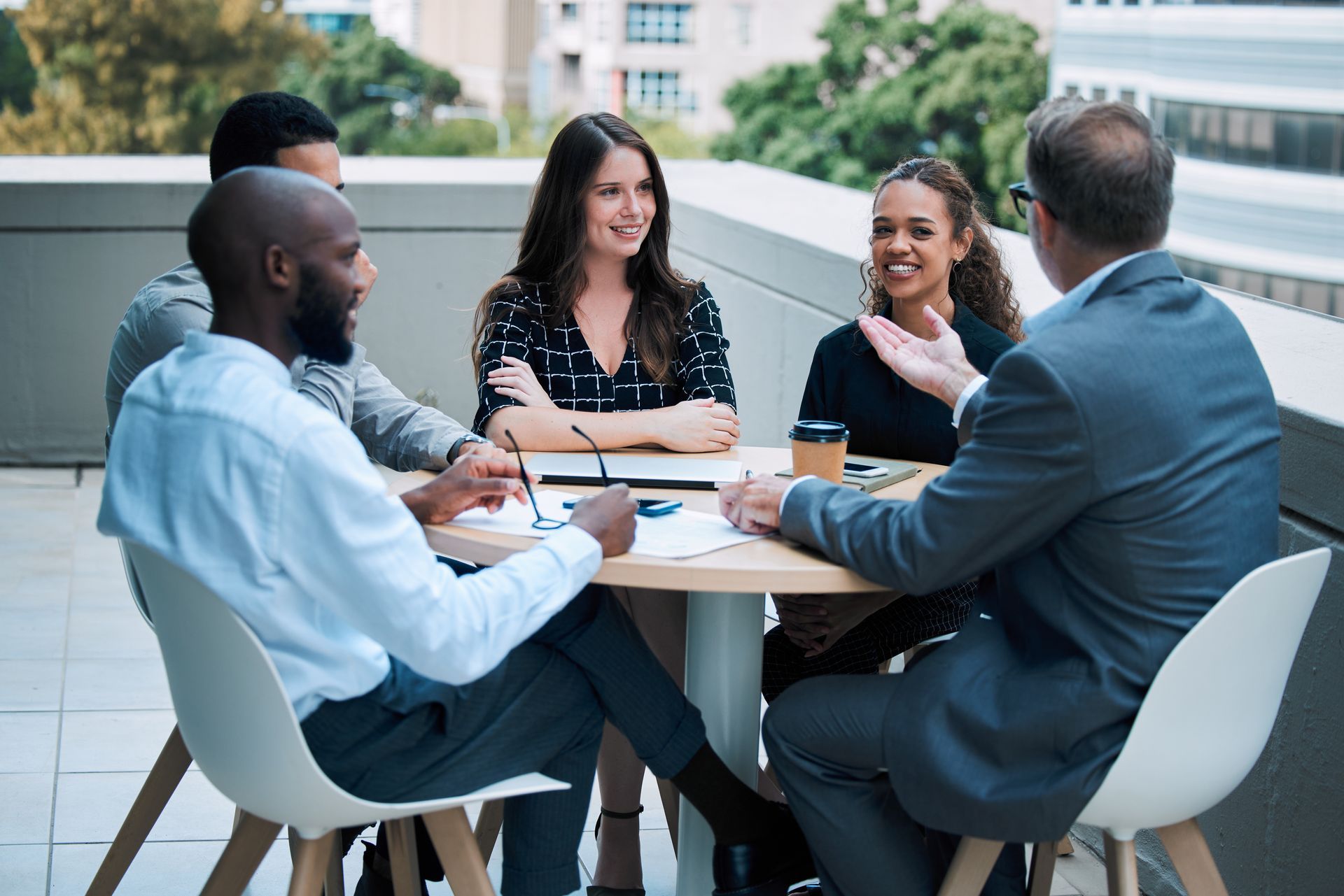 Four coworkers in suits and casual attire meet around a table outdoors, smiling and talking.