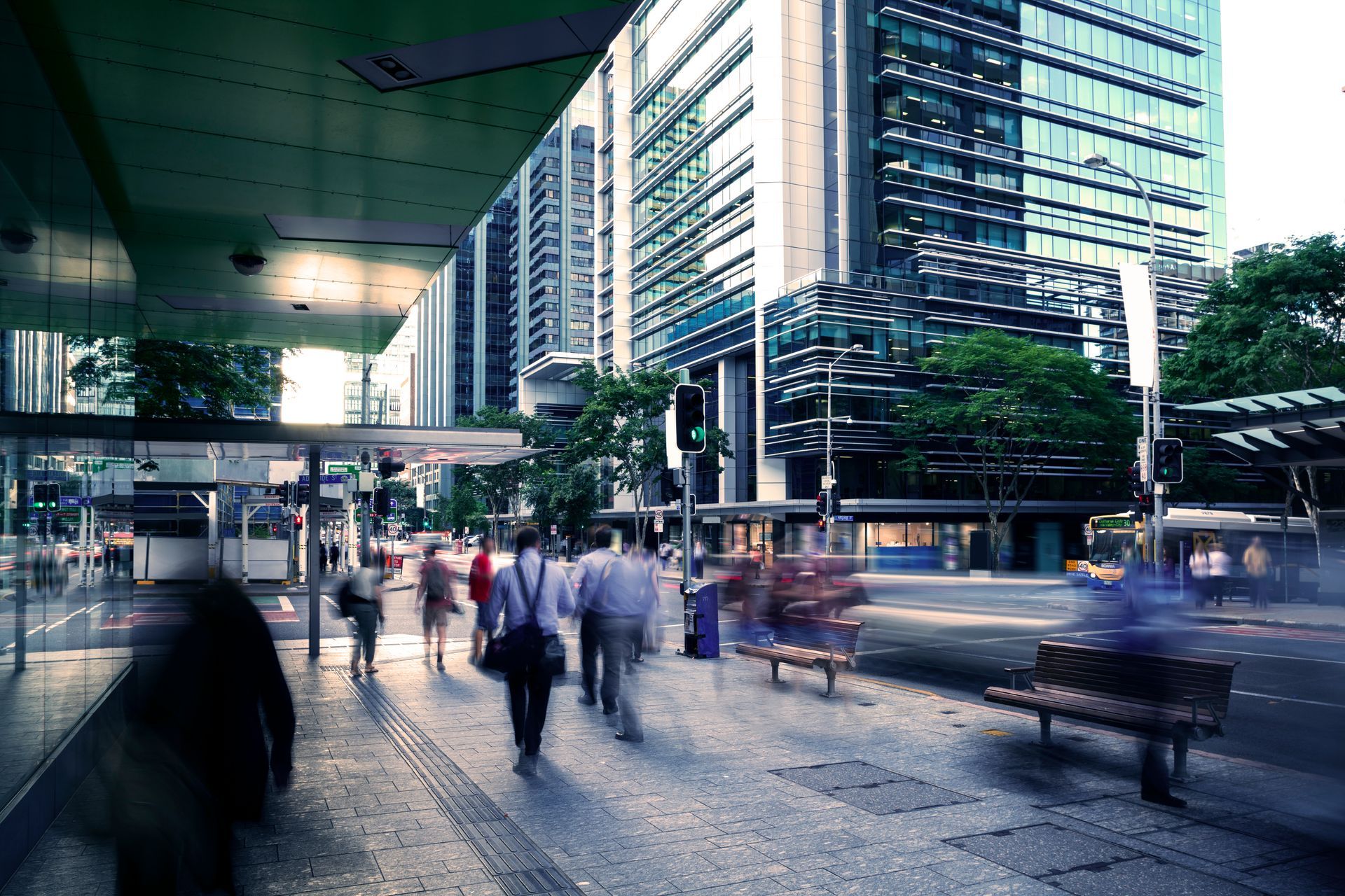 Busy city sidewalk with blurred pedestrians, benches, and tall glass office buildings in the background
