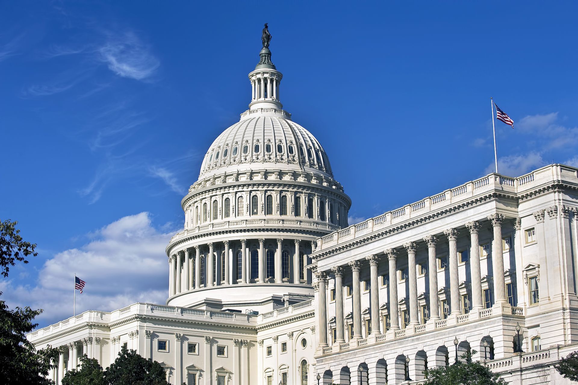 The U.S. Capitol building dome and neoclassical architecture against a bright blue sky with scattered clouds.