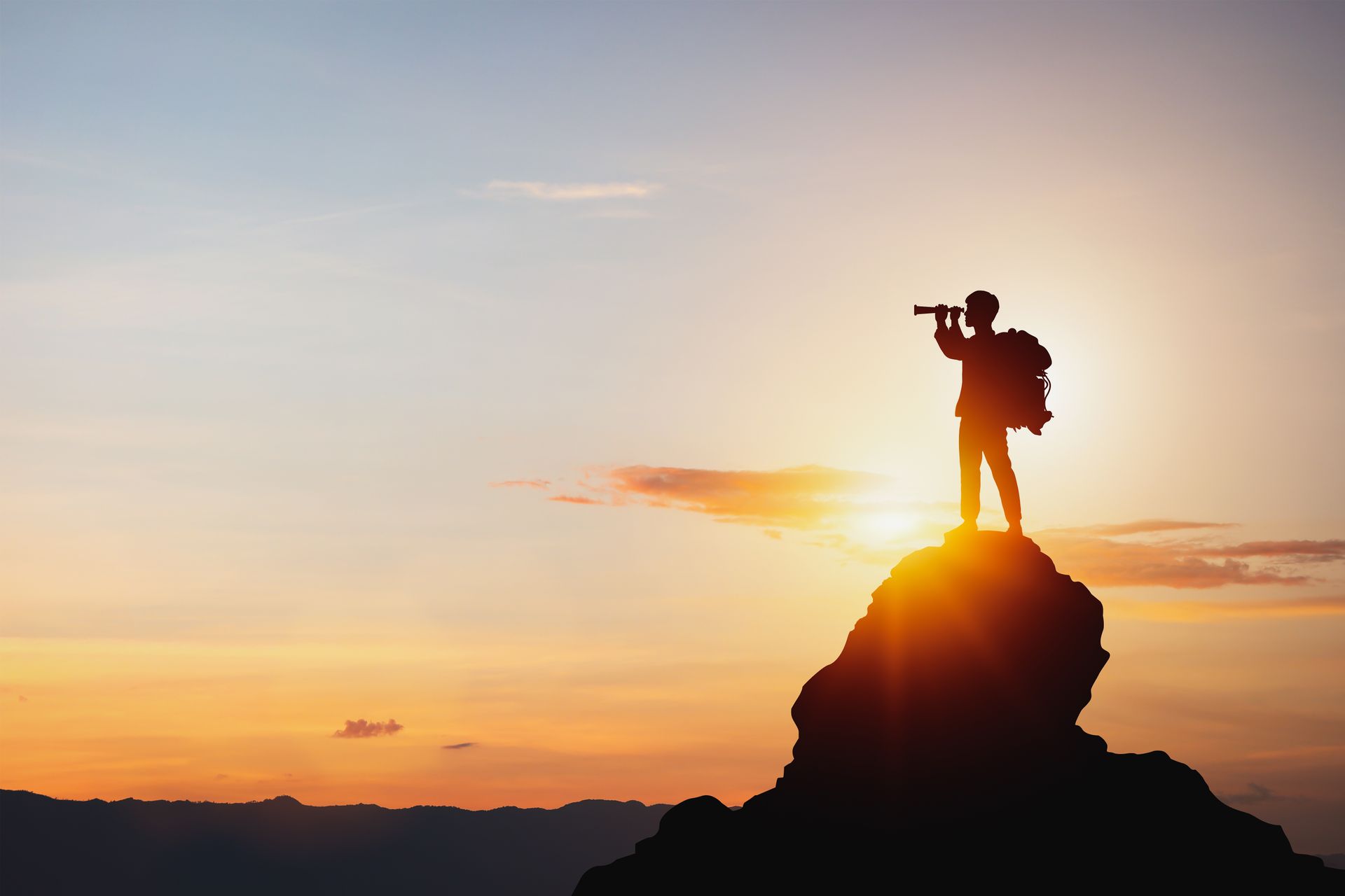 A man is standing on top of a mountain looking through a telescope at sunset.