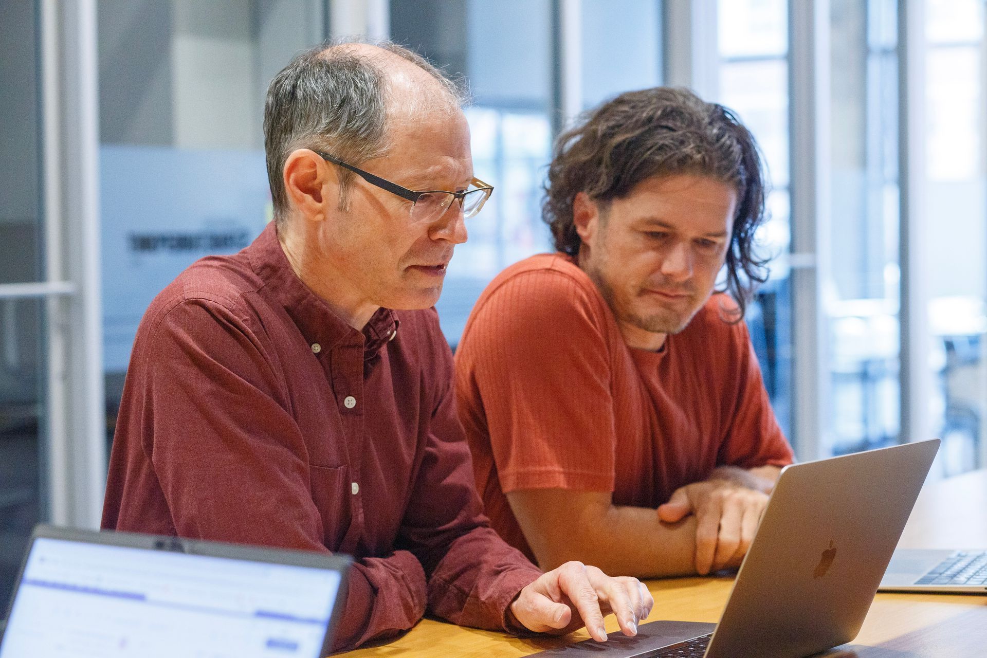 Two men are sitting at a table looking at a laptop computer.