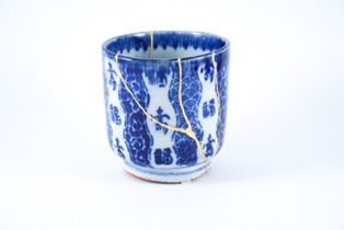 Xícara de cerâmica azul e branca, reparada com kintsugi dourado, sobre fundo branco.