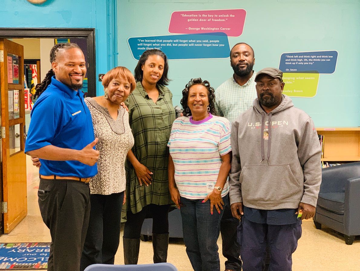 Six people stand together in a classroom, smiling toward the camera, in front of a wall with educational signage.