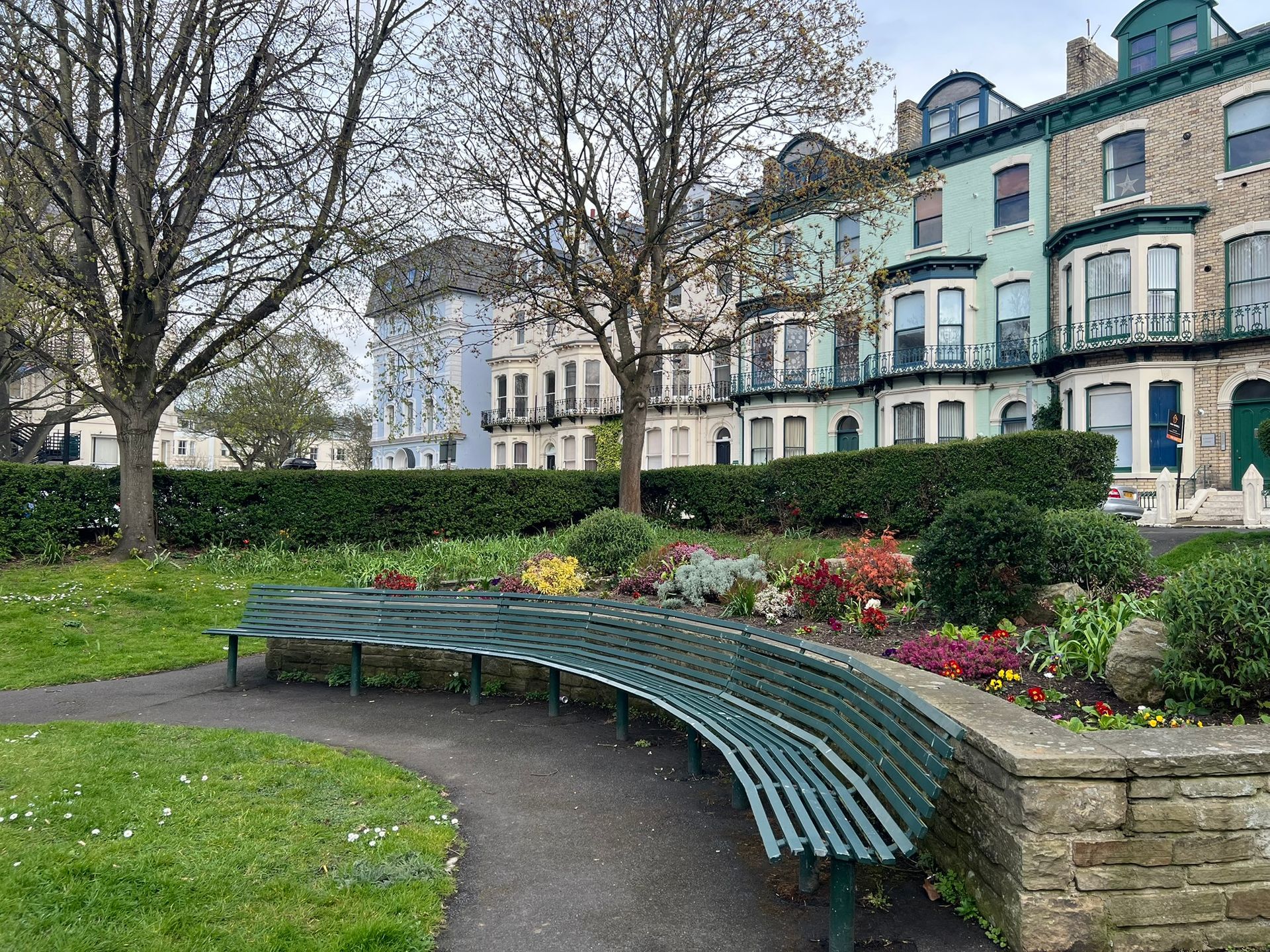 Curved green bench in a park garden beside flowers and historic buildings