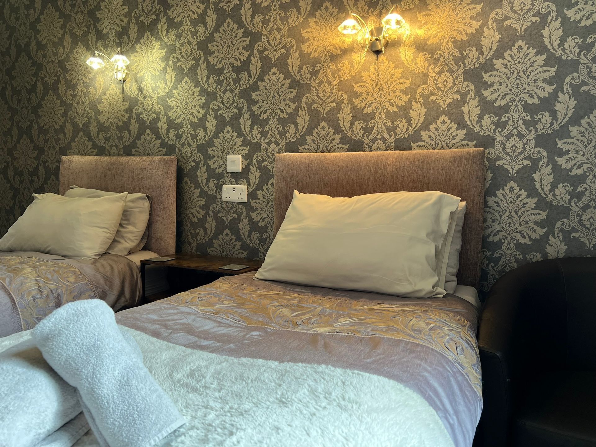 Two neatly made beds in a dimly lit bedroom with patterned wallpaper and wall sconces