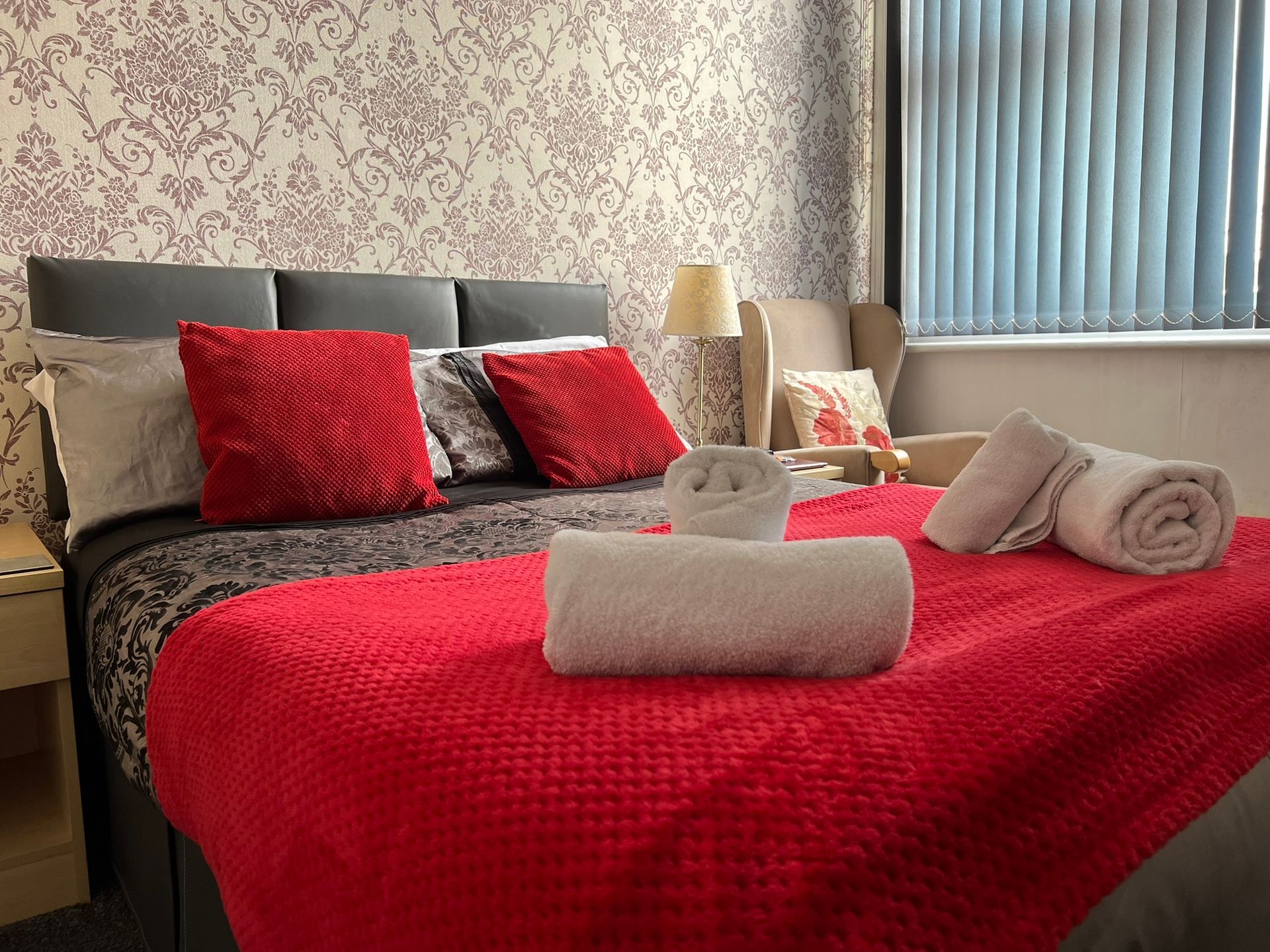 Bedroom with a red bedspread, gray pillows, rolled towels, and patterned wallpaper