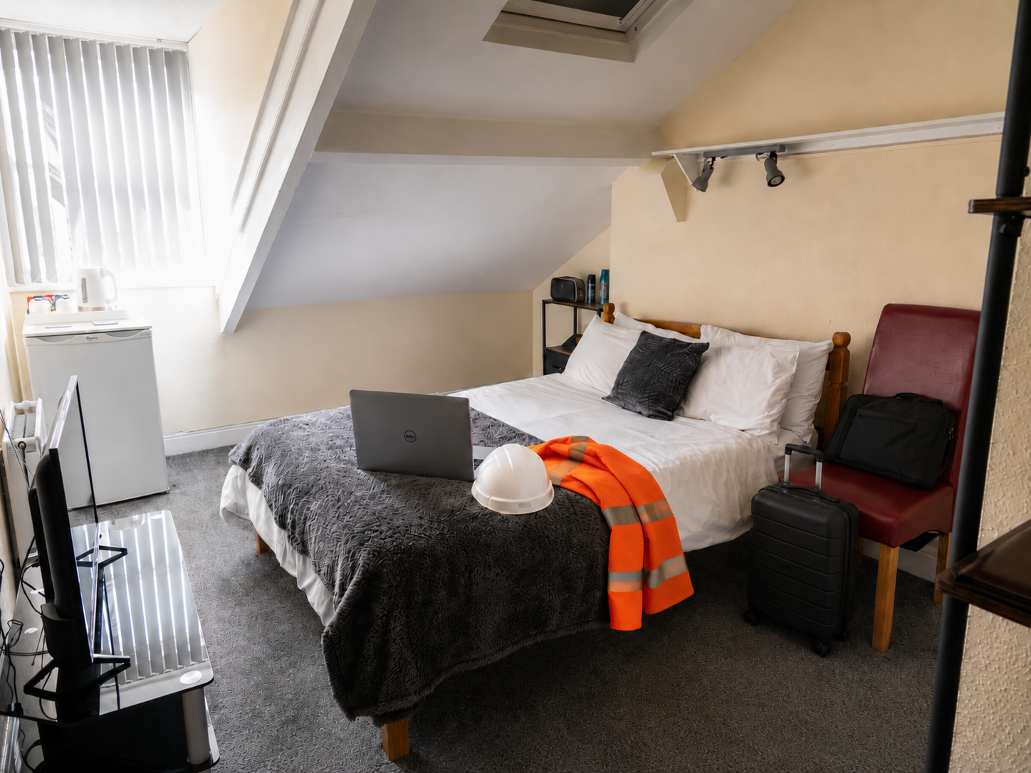 Contractors room showing laptop, hard hat and hi-viz on a double bed, small suitcase and toiletries