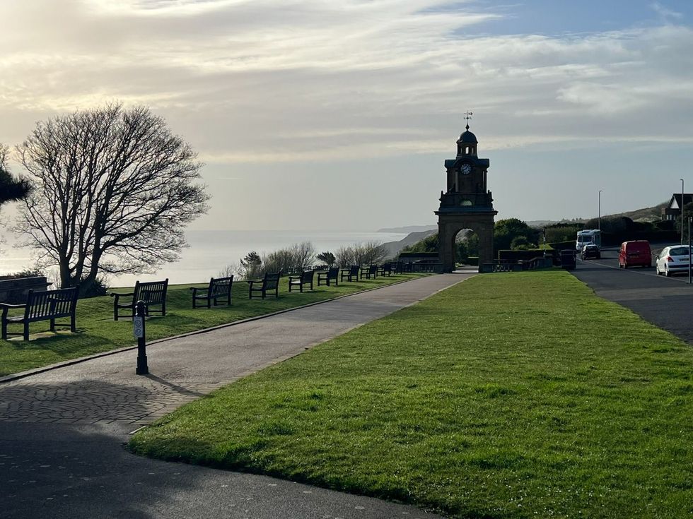 Path leading to a stone monument tower beside a grassy hill, with a tree and parked cars under a cloudy sky