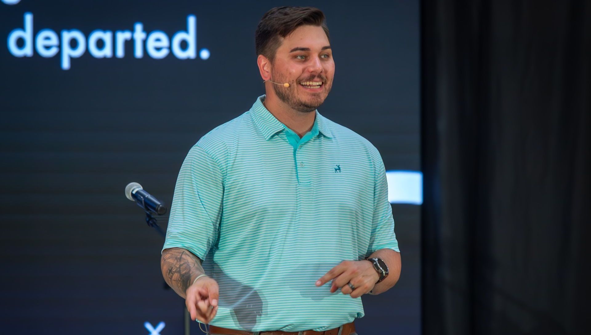 Man in a light teal polo speaks on stage beside a microphone, with “departed.” on the screen behind him.