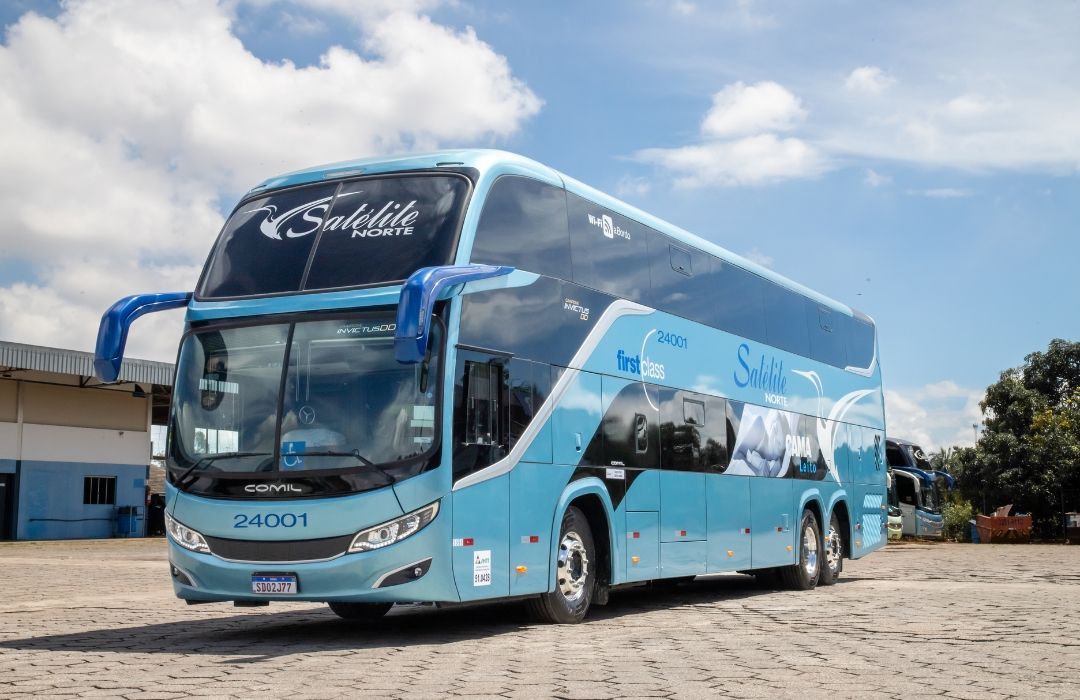Ônibus de luxo azul-claro e preto estacionado em um terreno arenoso sob um céu nublado.