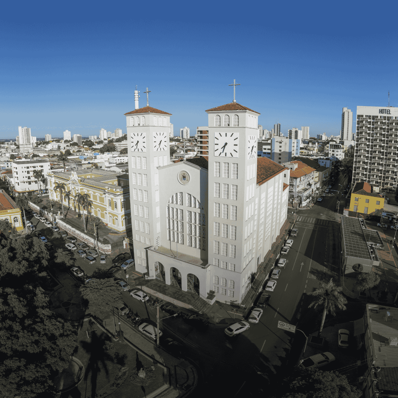 A imagem apresenta uma vista aérea de uma área urbana, tendo como destaque uma grande igreja de arquitetura imponente no centro da composição. O edifício é branco, possui duas torres simétricas com relógios em suas fachadas e cruzes no topo, além de telhados com telhas avermelhadas. A entrada principal conta com arcos e uma ampla escadaria.