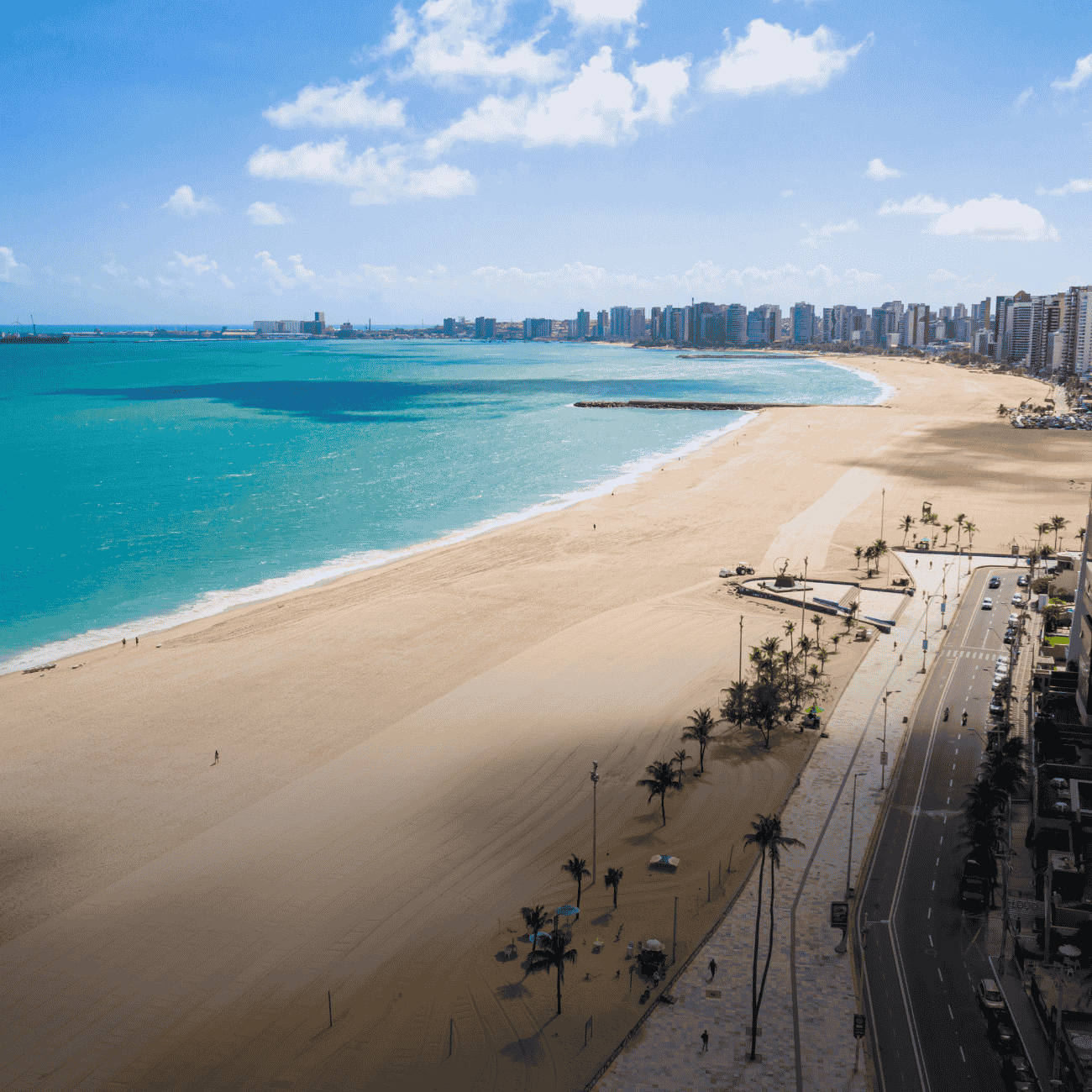 A imagem mostra uma vista aérea de uma extensa praia urbana em um dia ensolarado, com mar em tons de azul-turquesa e uma larga faixa de areia clara. Ao fundo, uma longa fileira de edifícios à beira-mar compõe o horizonte da cidade, enquanto a avenida litorânea, ladeada por coqueiros, acompanha toda a orla.
