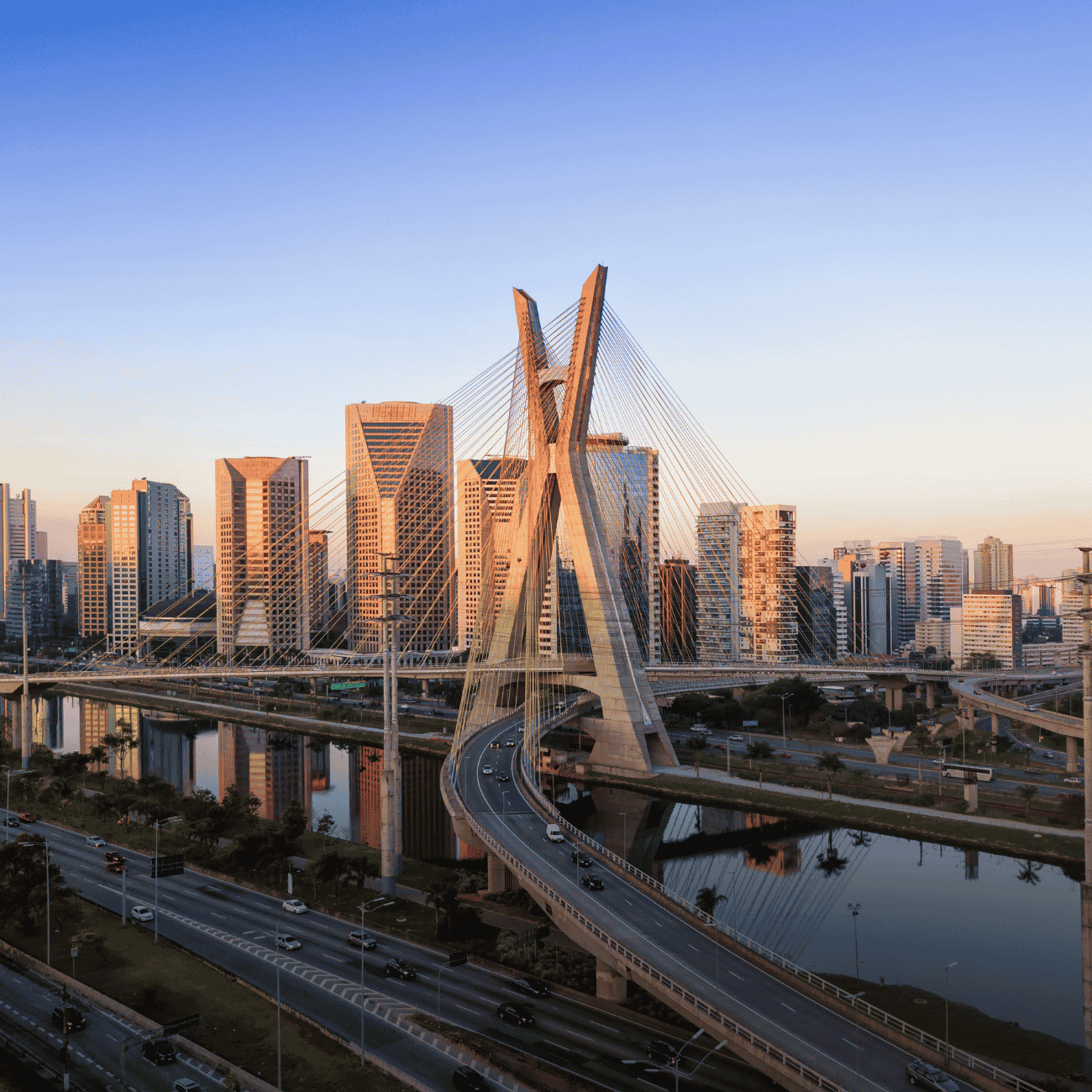 A imagem apresenta uma vista panorâmica da cidade de São Paulo ao entardecer, destacando a icônica Ponte Estaiada sobre o Rio Pinheiros.
Os prédios modernos da região empresarial refletem a luz dourada do pôr do sol, criando uma atmosfera urbana sofisticada.