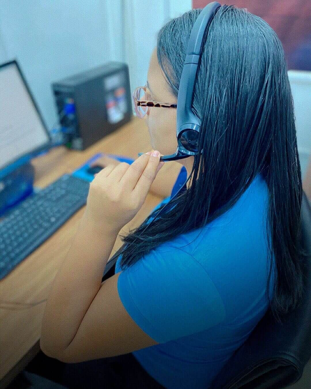 Uma pessoa vestindo uma camisa azul clara e um fone de ouvido, sorrindo enquanto ajusta o microfone com a mão.