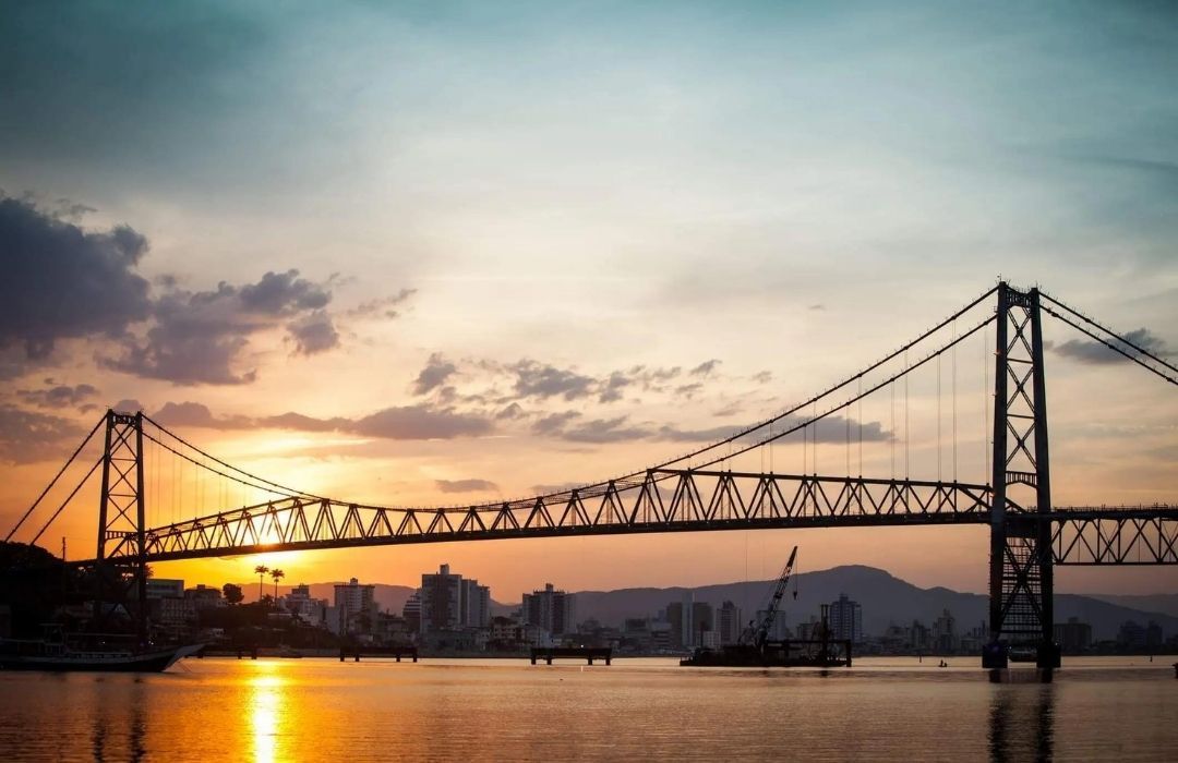 Ponte suspensa ao pôr do sol sobre águas calmas, com o horizonte da cidade e reflexos alaranjados em tons quentes.