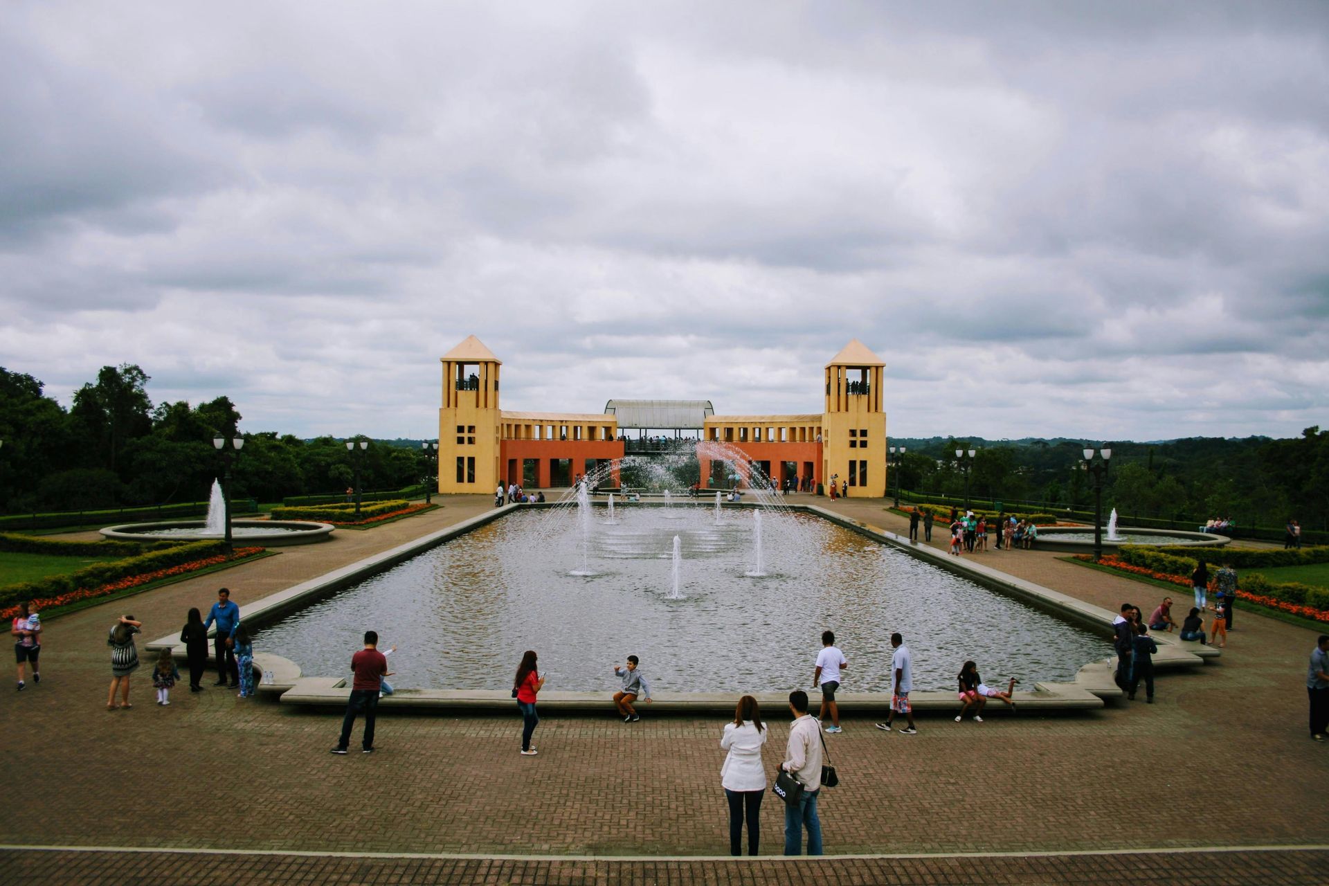 Uma grande fonte retangular com múltiplos jatos de água em frente a um prédio amarelo com duas torres, sob um céu nublado.