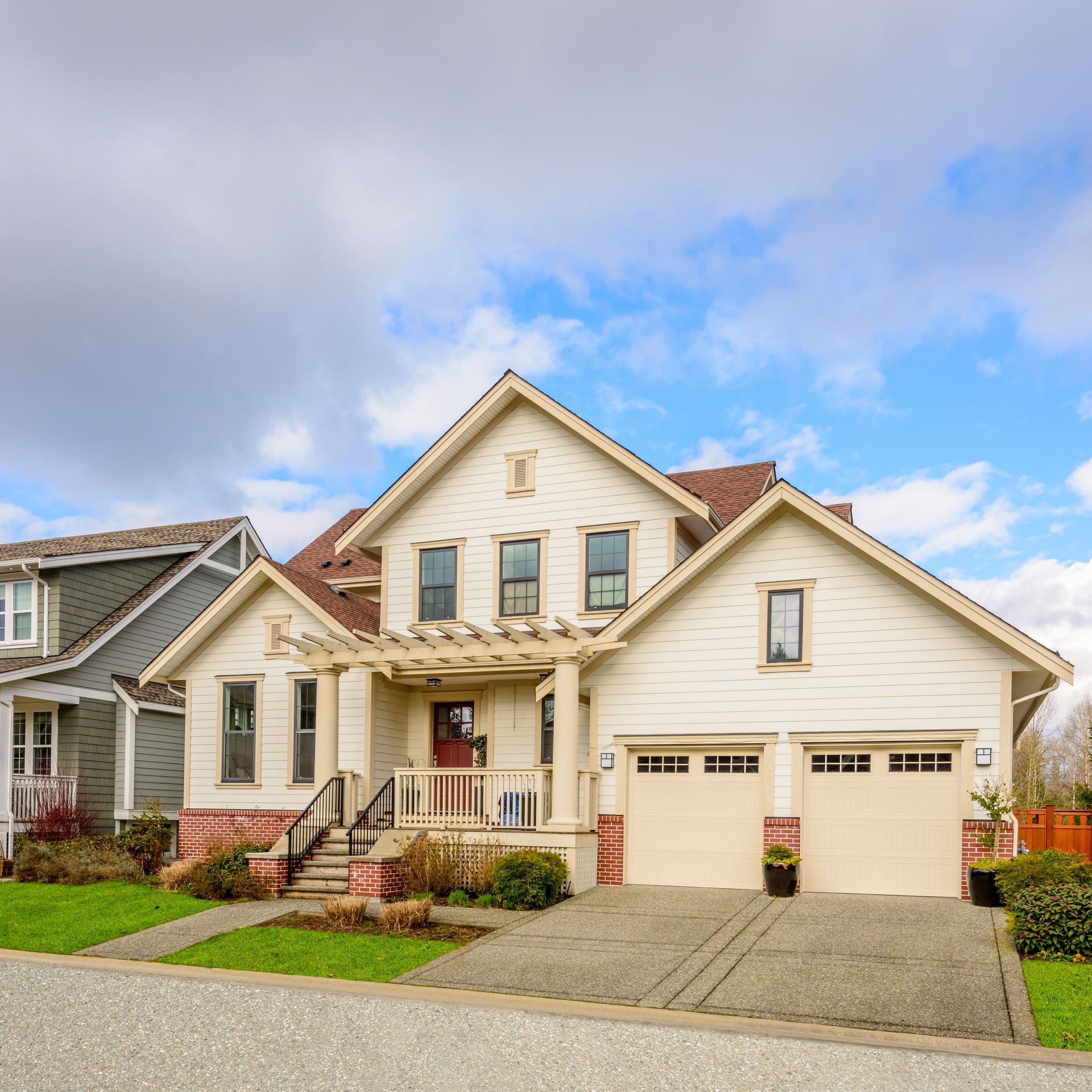 Beige two-story house with a two-car garage, red door, and pergola. Blue sky with clouds. Beige two-story house with a two-car garage, red door, and pergola. Blue sky with clouds.