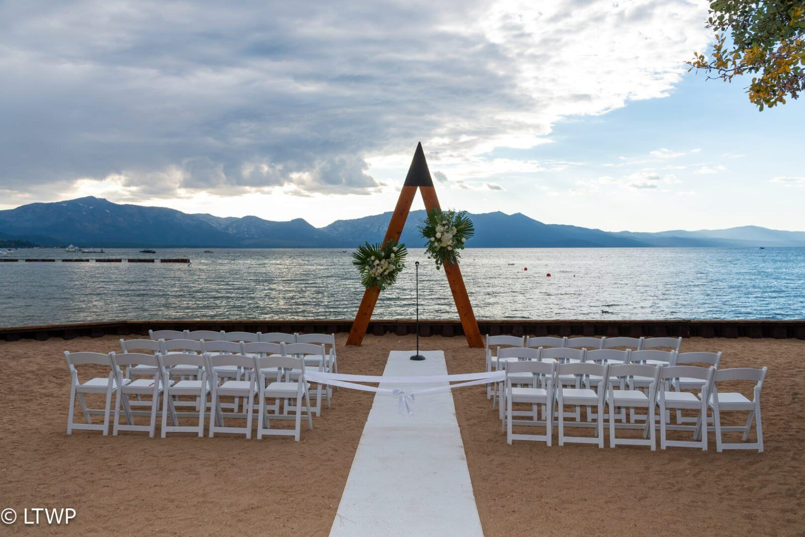 Wedding ceremony setup on a sandy beach with white chairs, wooden arch, and ocean view.