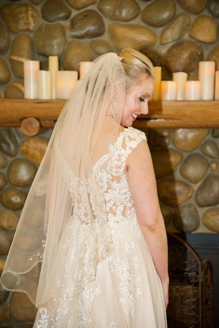 Bride in lace wedding dress, veil, smiles near a stone fireplace with candles.