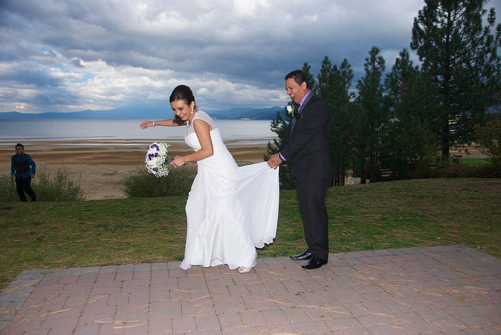 Bride throws bouquet, groom helps with dress. Lakeside setting, cloudy sky.