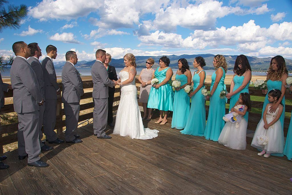 Wedding ceremony on wooden deck: bride, groom, bridesmaids in turquoise, groomsmen in gray, mountains in background.