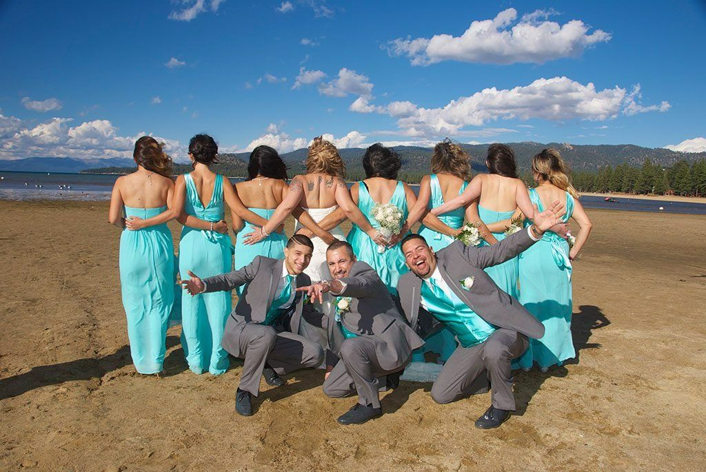 Wedding party on beach, bridesmaids in teal dresses, groomsmen in gray suits, posing playfully.