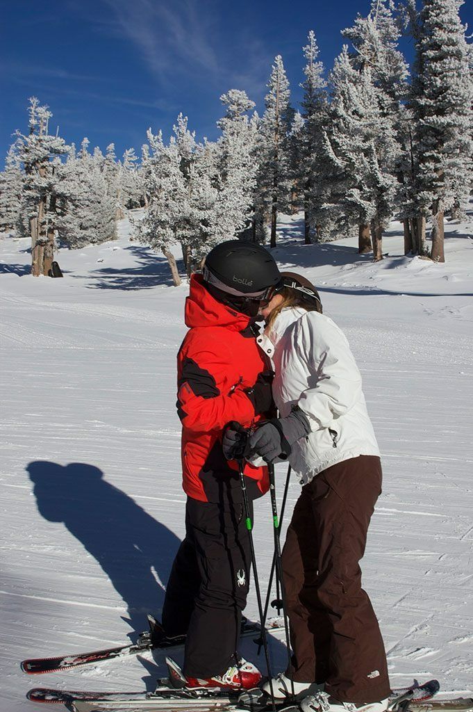 Couple kissing on skis in a snowy mountain landscape, wearing winter coats and helmets.