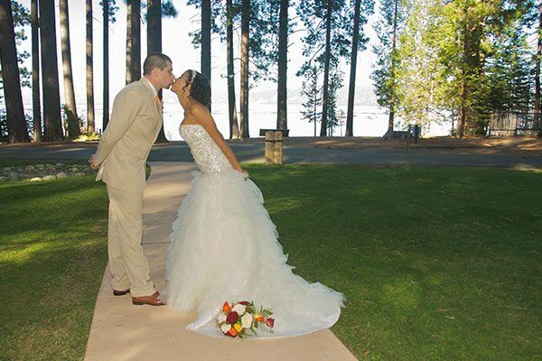 Groom in beige suit kisses bride in white gown outdoors on grass path; bouquet at her feet.