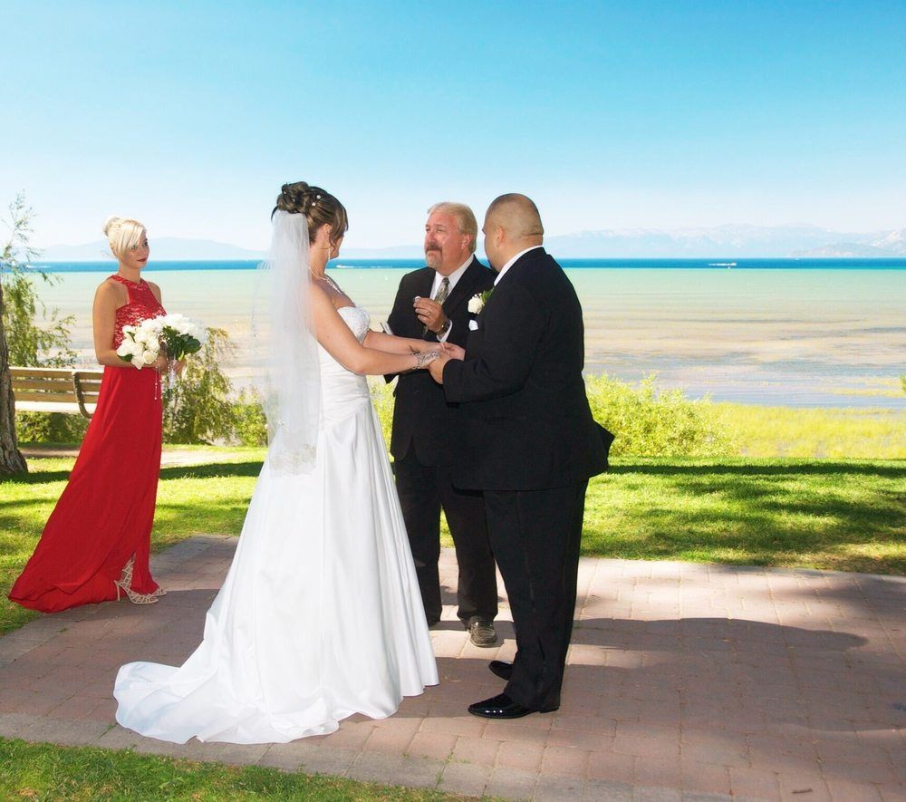 Wedding ceremony on a sunny day by the water. Bride and groom exchange vows, officiant present, bridesmaid in red.