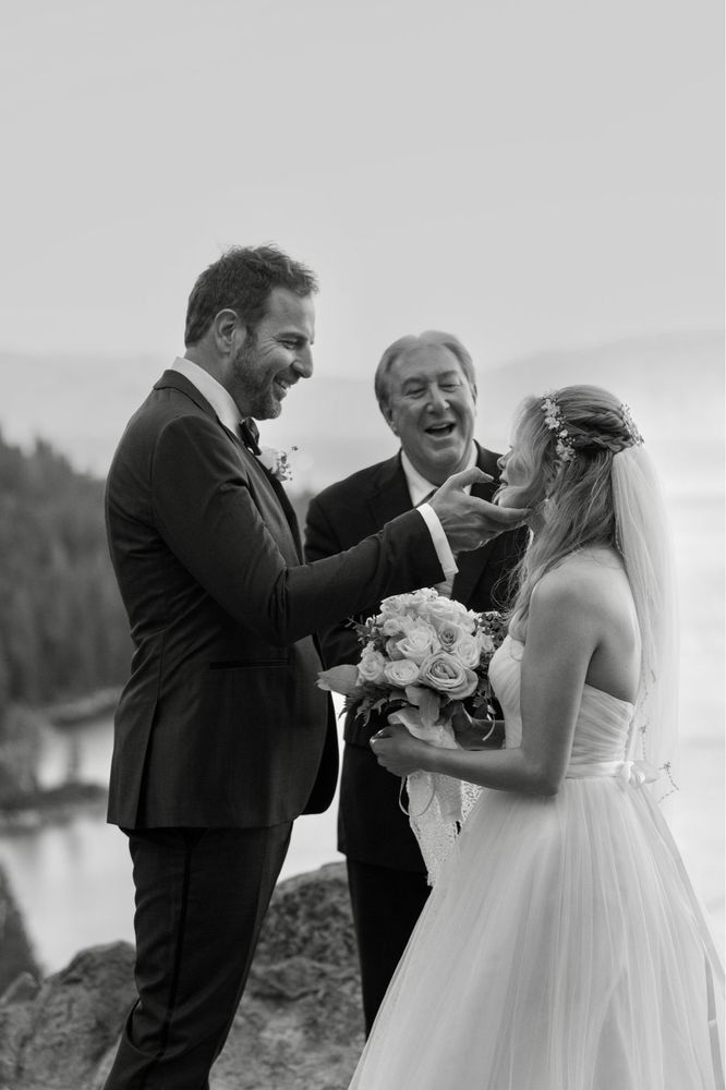 Groom touches bride's face during outdoor wedding ceremony; officiant smiles.