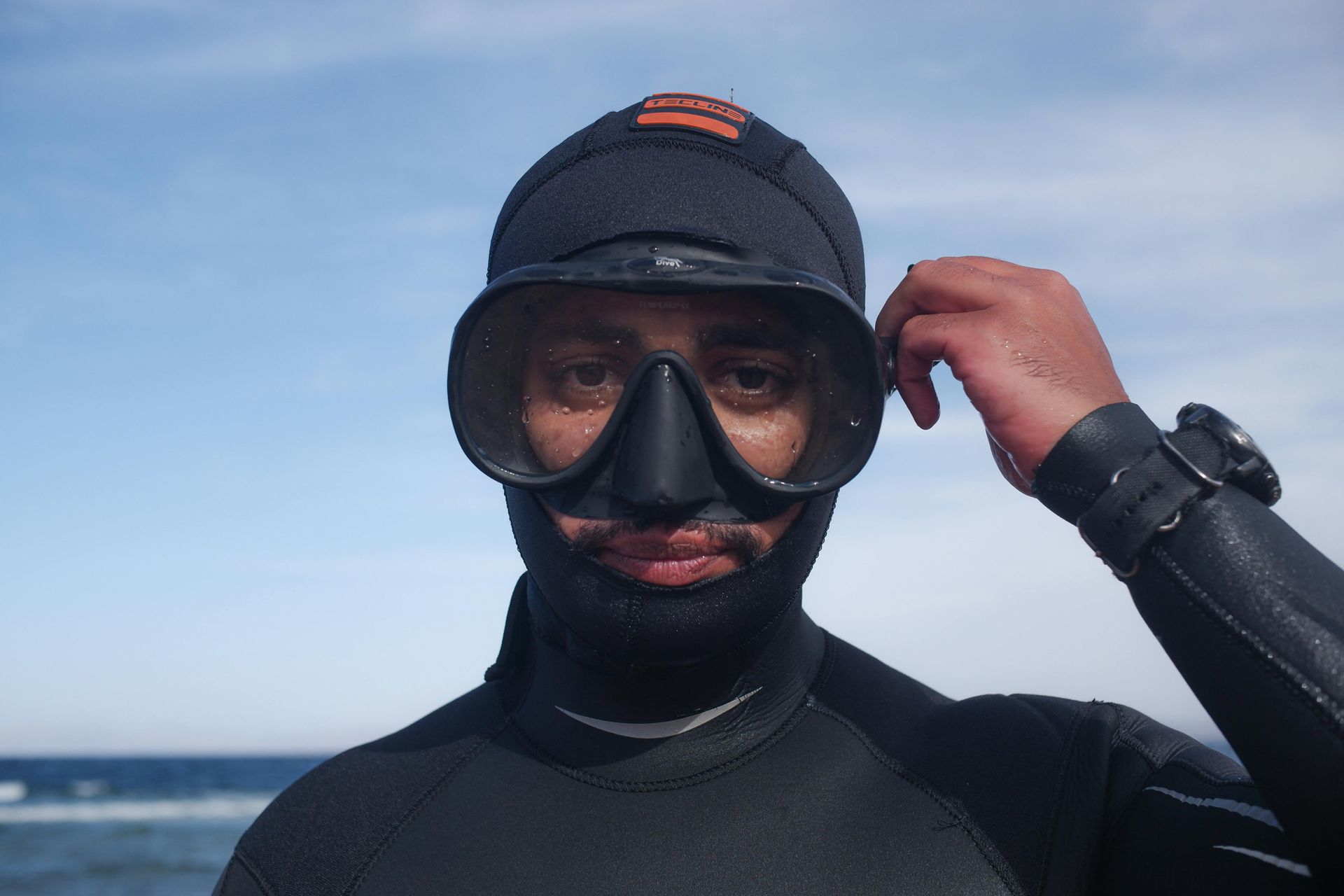 Person in a black wetsuit and goggles adjusts their cap by the ocean.