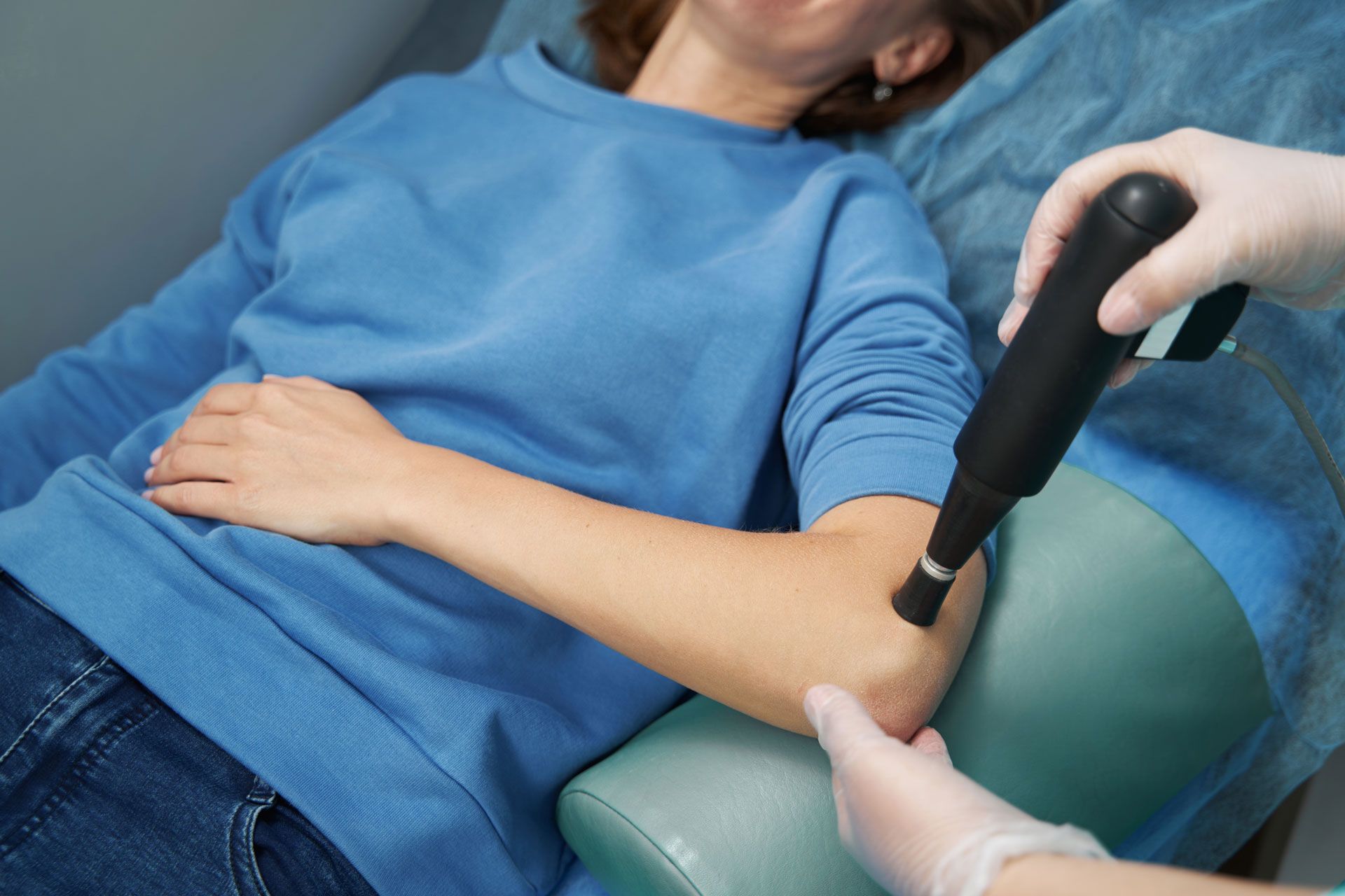 A person receives medical treatment on their elbow with a handheld device; setting is a clinic.