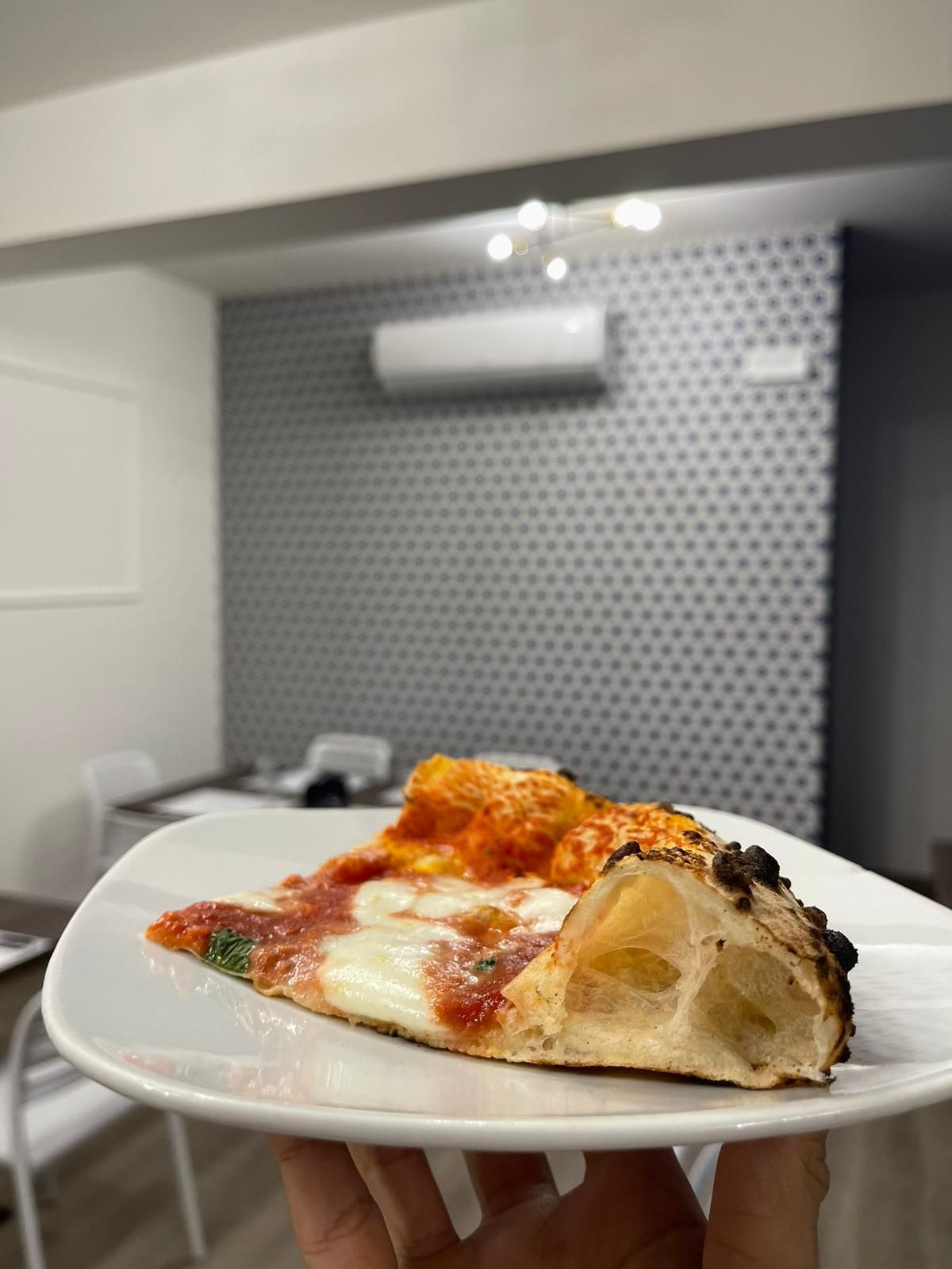 In una sala da pranzo, una mano regge un piatto con una fetta di pizza cotta nel forno a legna, con crosta croccante.