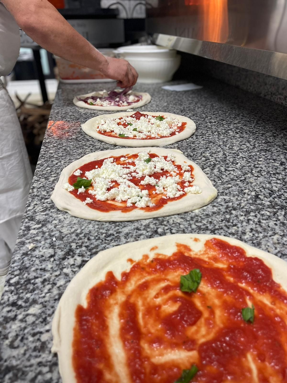 In una cucina, una persona compone una fila di pizze con salsa di pomodoro, formaggio e basilico su un piano di lavoro.