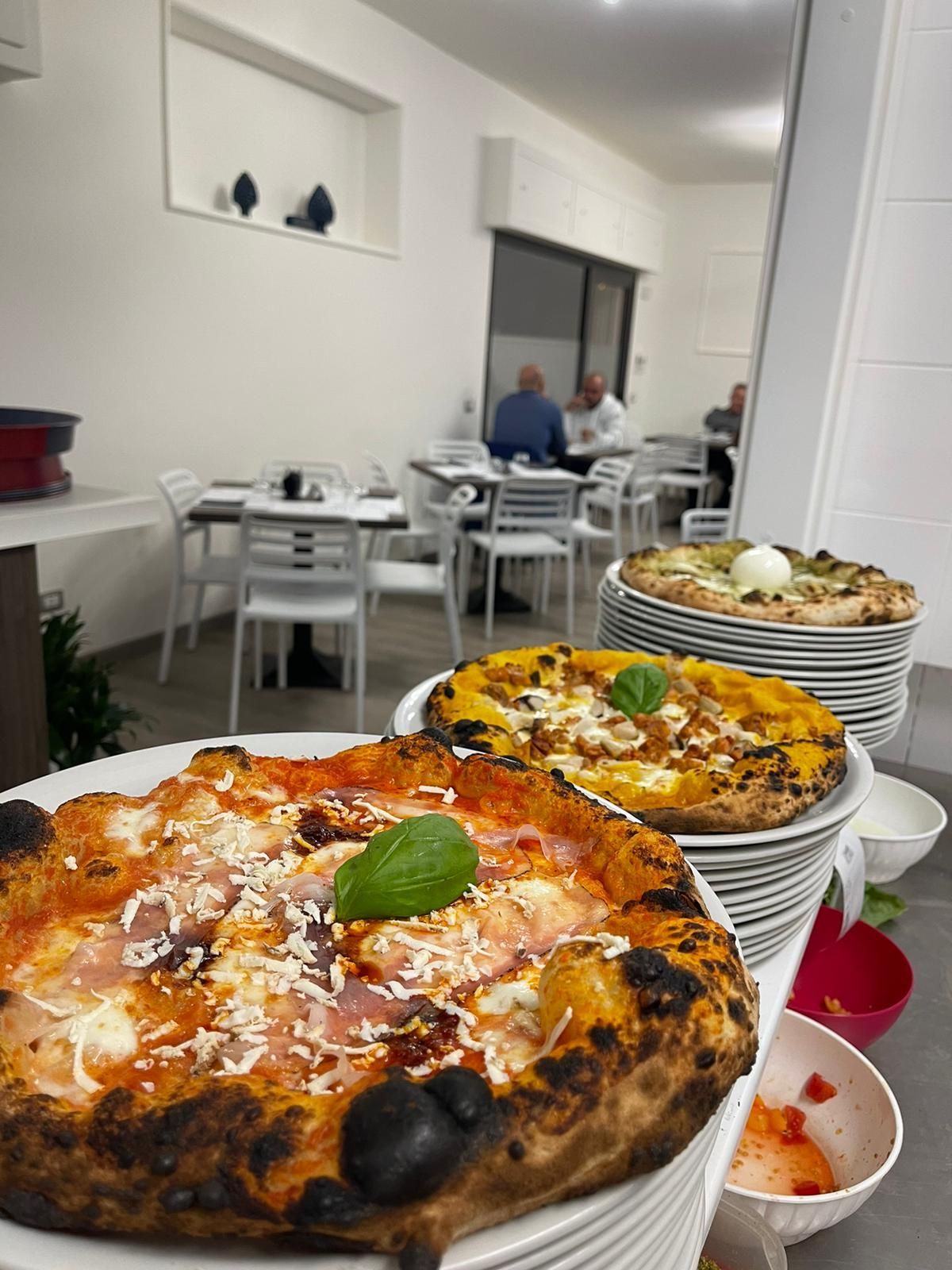 In una cucina di ristorante con tavoli, si vedono pile di pizze fresche cotte nel forno a legna, con vari condimenti.