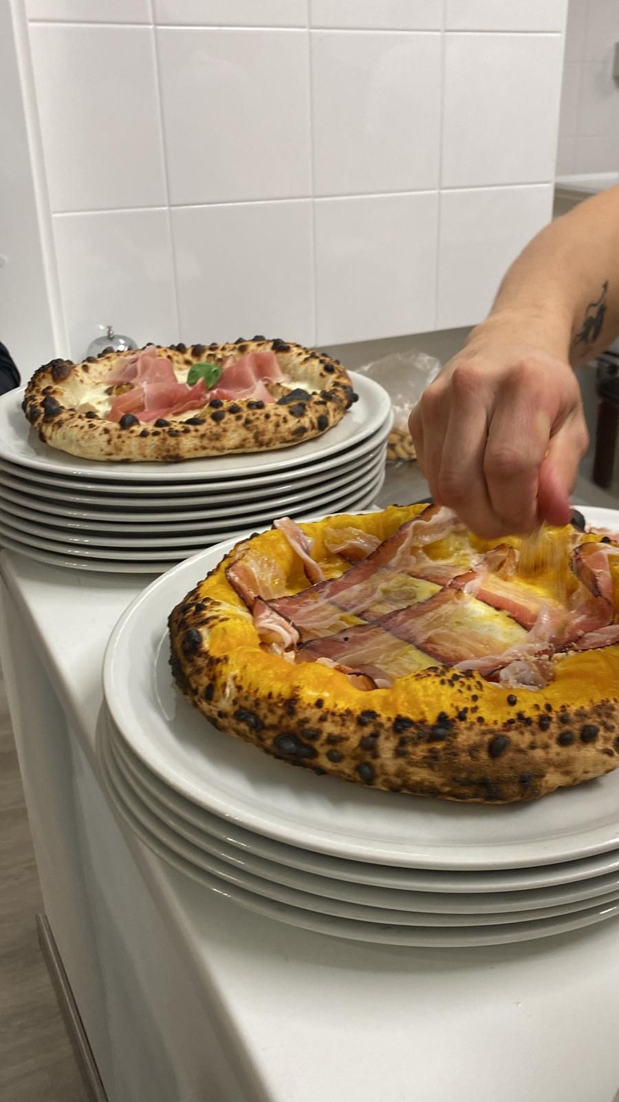 Una persona guarnisce una pizza con prosciutto e salsa, mentre un'altra pizza è appoggiata su una pila di piatti lì vicino.