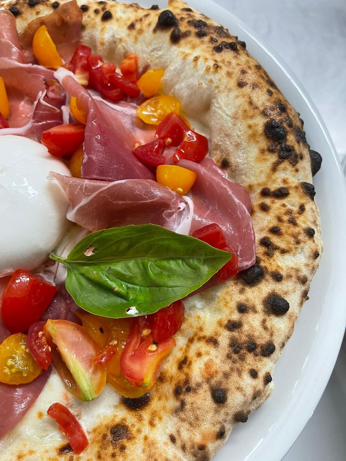 Primo piano di una pizza condita con burrata fresca, prosciutto crudo a fette sottili, pomodorini gialli e rossi e basilico.