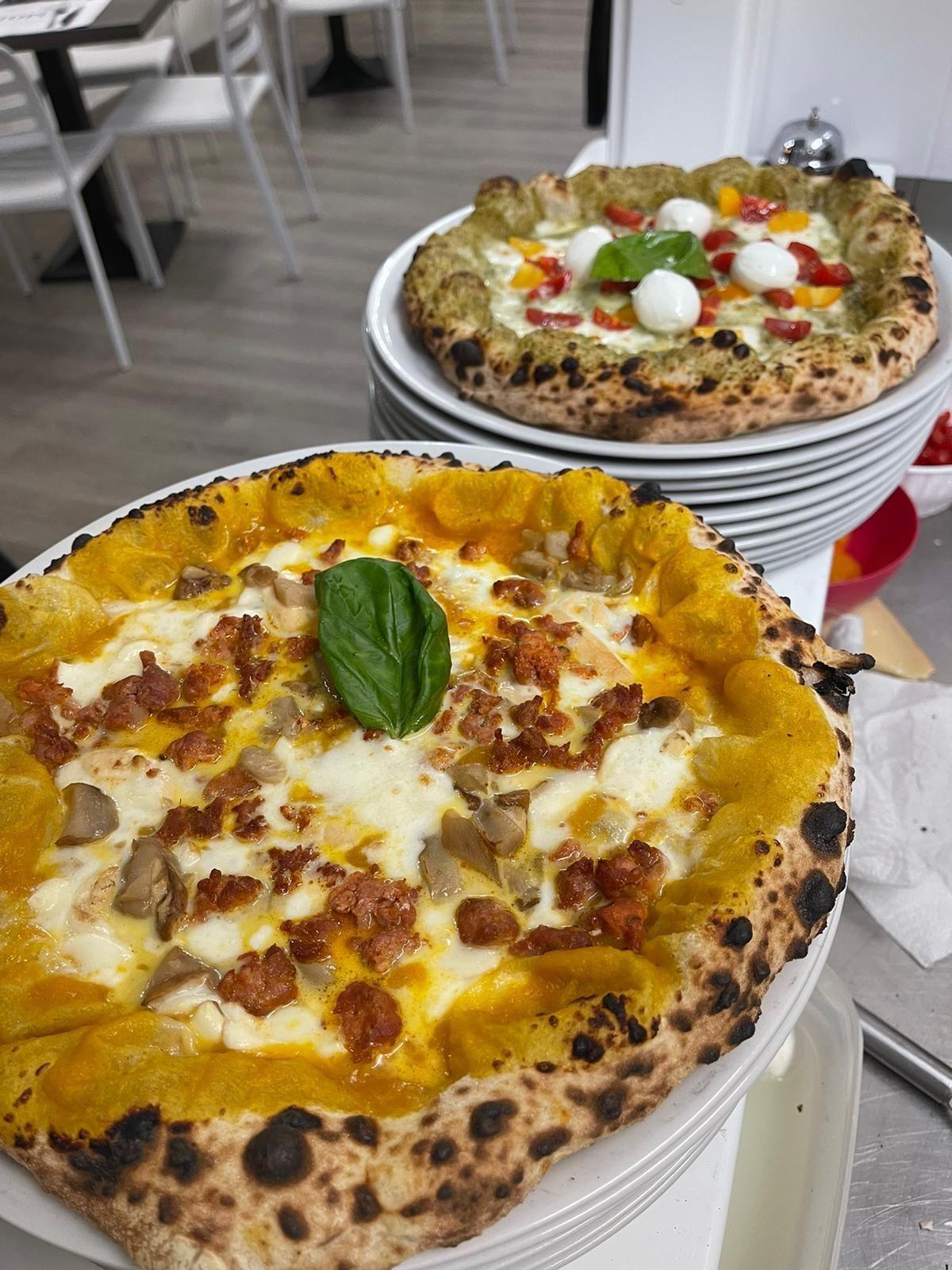 Due pizze su piatti bianchi: una con base di zucca dorata, funghi e pancetta; l'altra con pesto, pomodori e formaggio.