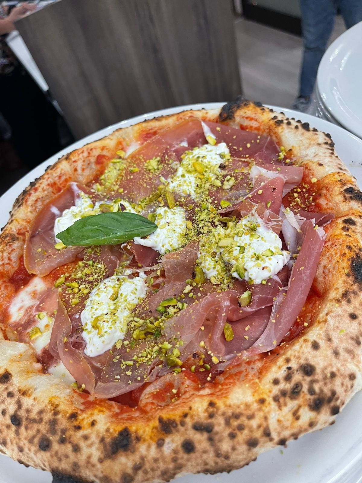 Una pizza con prosciutto crudo, fiocchi di formaggio bianco e pistacchi tritati, guarnita con una singola foglia di basilico.