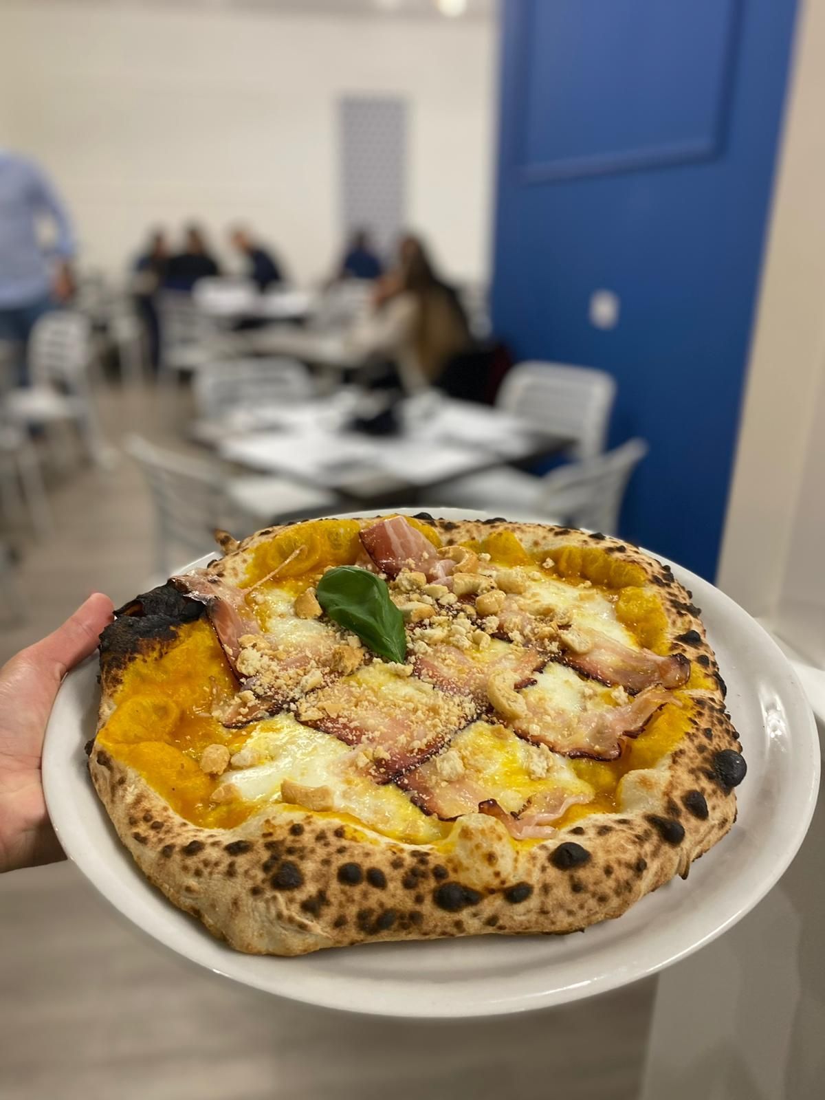 Una persona tiene in mano una pizza condita con zucca, formaggio fuso, salumi e noci in un ristorante con i tavoli sfocati.