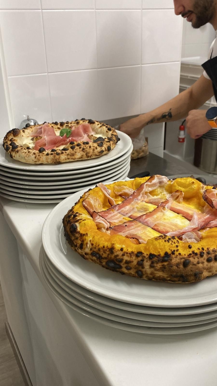 In cucina, una persona prepara due pizze appena sfornate su pile di piatti bianchi.