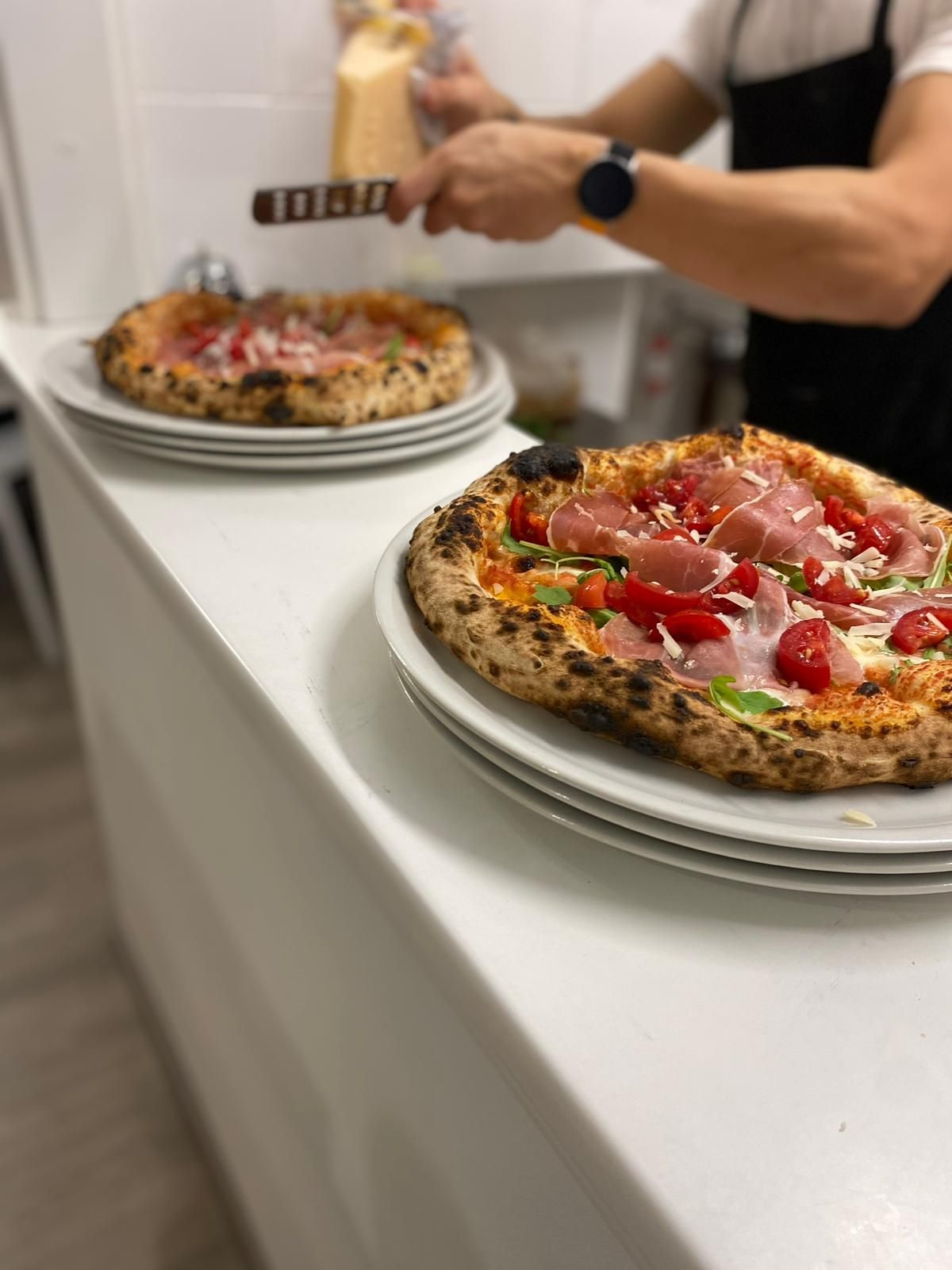 In cucina, una persona grattugia del formaggio su una pizza appena sfornata, mentre un'altra pizza è appoggiata lì vicino.