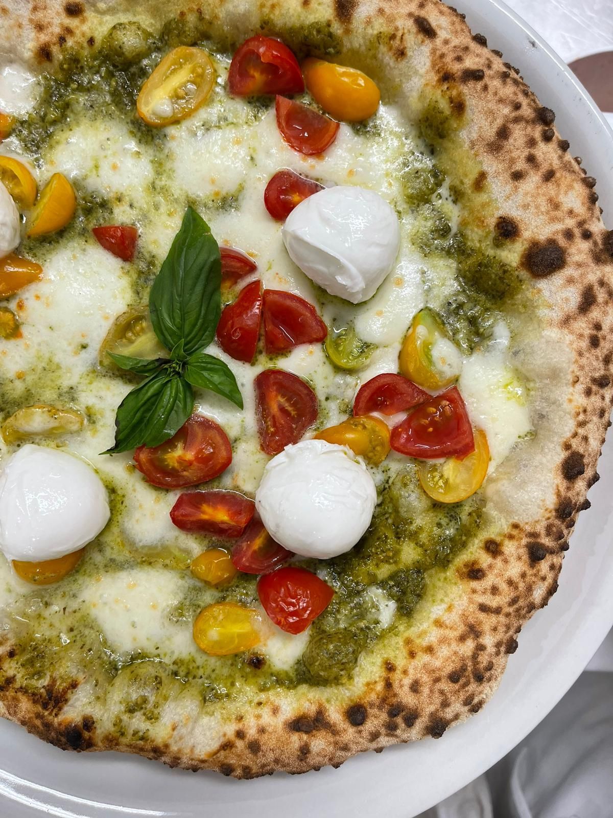 Una pizza al pesto con mozzarelline, pomodorini rossi e gialli e basilico fresco, servita su un piatto bianco.